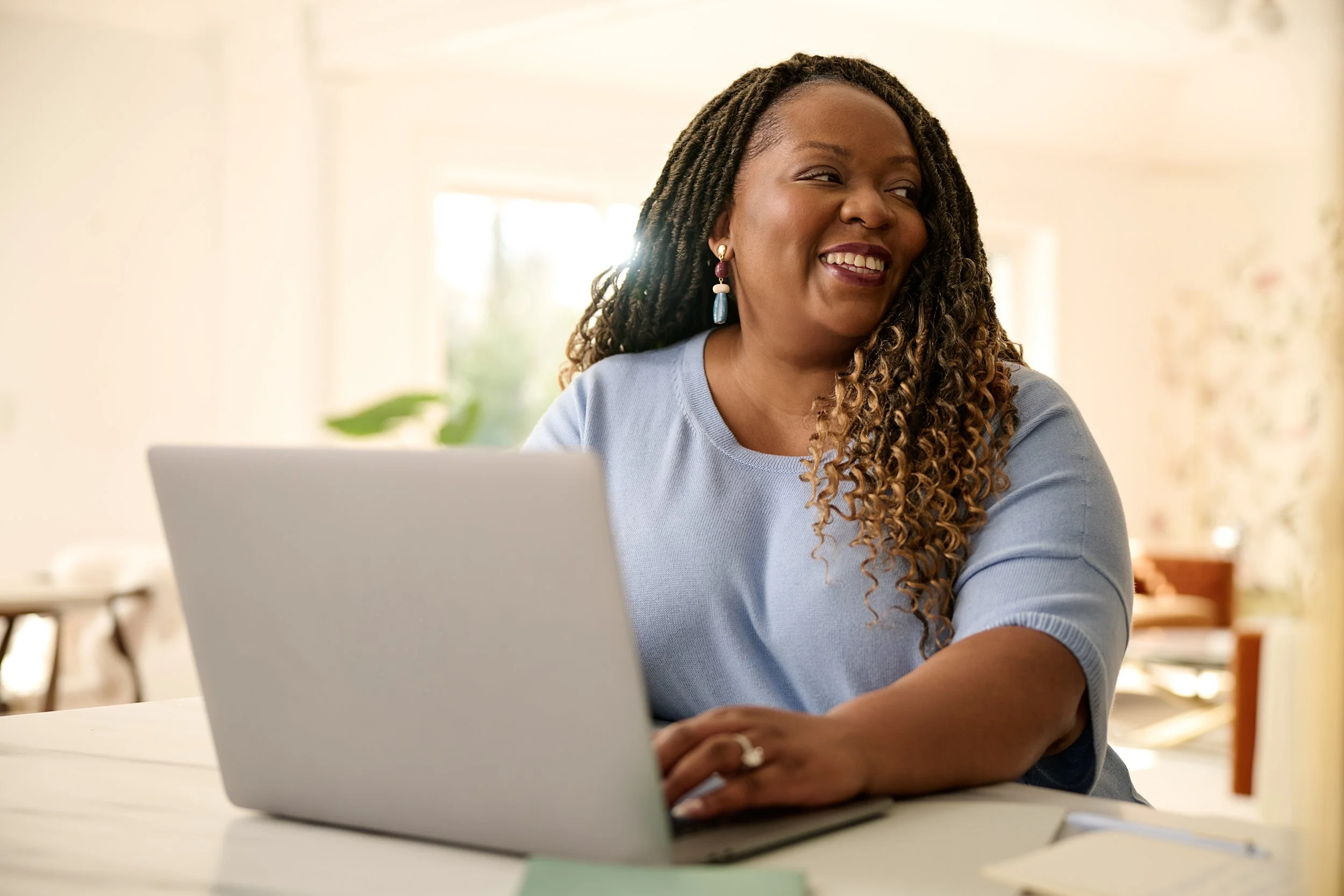 A woman with long, curly hair wearing earrings and a blue top sitting at a desk with a laptop, smiling and looking to her left, in a well-lit room with a window in the background.