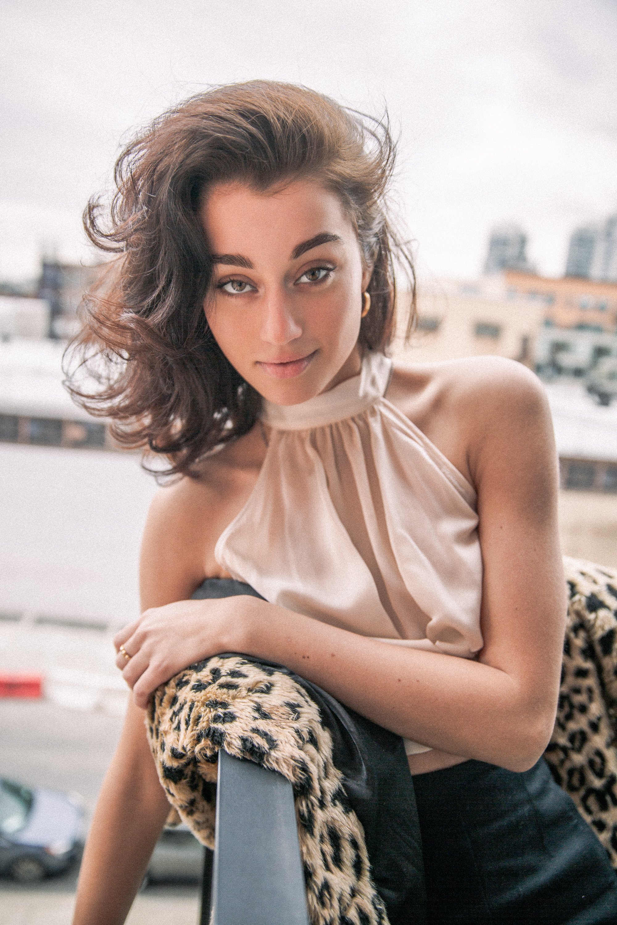 A woman with wavy brown hair and light eye makeup posing outdoors on a cloudy day, wearing a beige top with a keyhole neckline and a leopard print jacket draped over her arm.