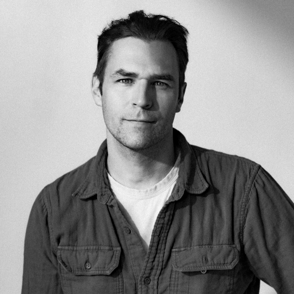 Black and white portrait of a man with dark hair, wearing a button-up shirt over a T-shirt, looking into the camera with a slight smile.