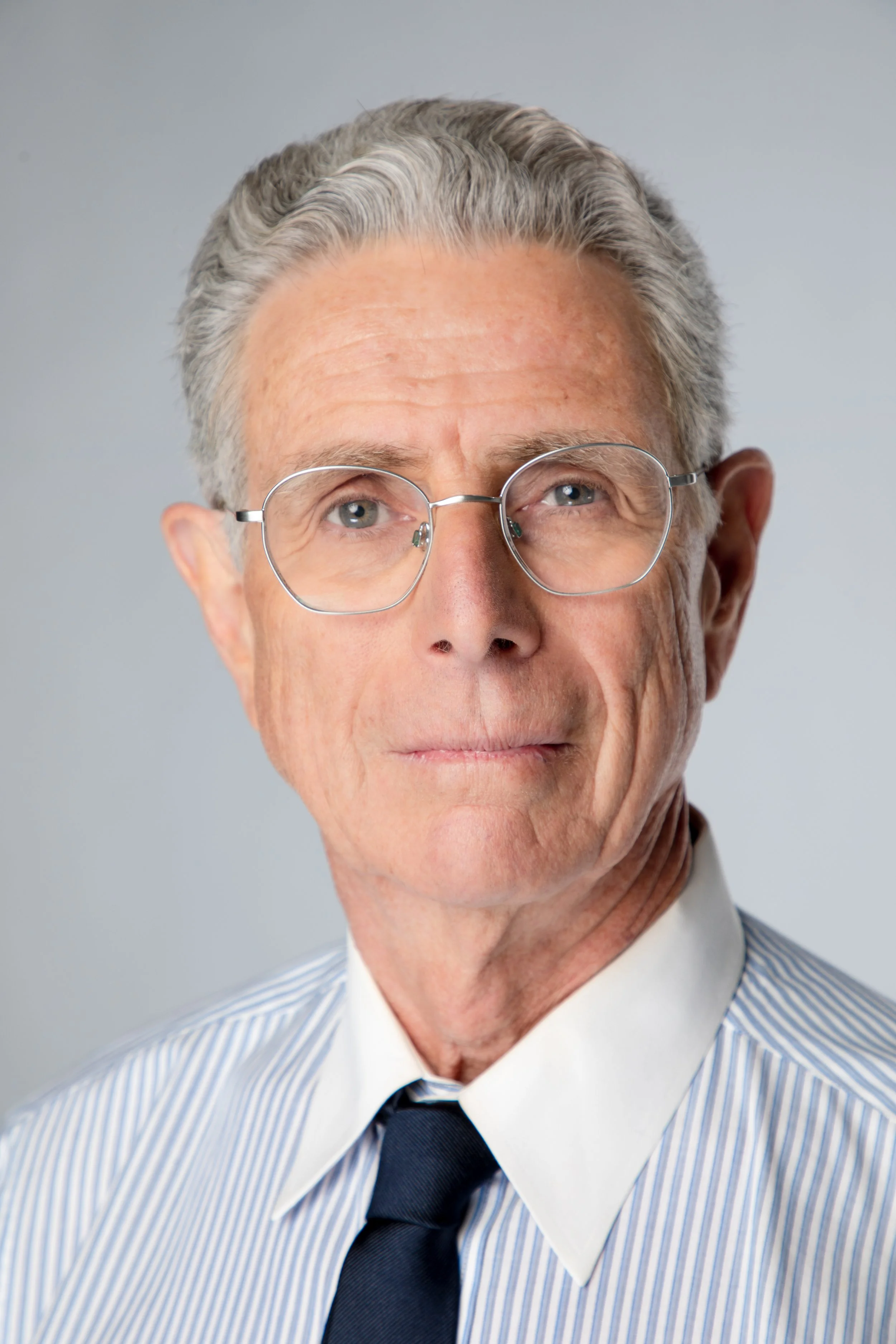Close-up of an elderly man with gray hair, wearing round glasses, a striped dress shirt, dark tie, and a white collar. He has a serious expression and is looking slightly to the side against a plain light background.