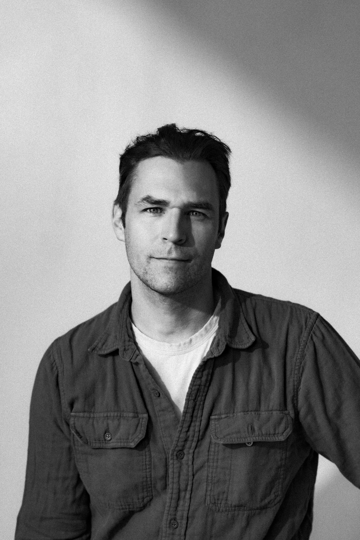 A black-and-white portrait of a young man with dark hair styled back, wearing a button-up shirt over a t-shirt, standing against a plain background with shadows cast on the wall.