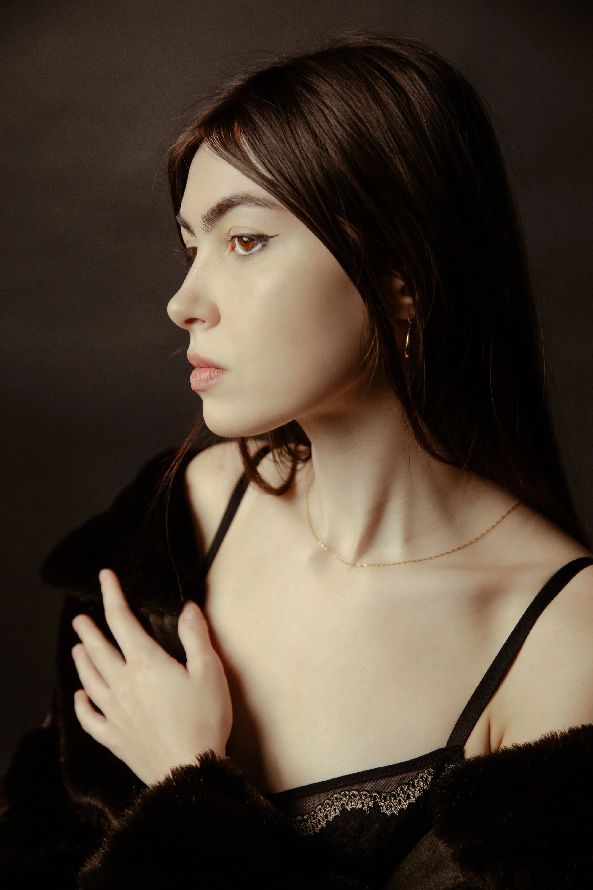 A young woman with brown hair, wearing black lace lingerie and a black furry robe, looking to the side with her hand on her chest, against a dark background.