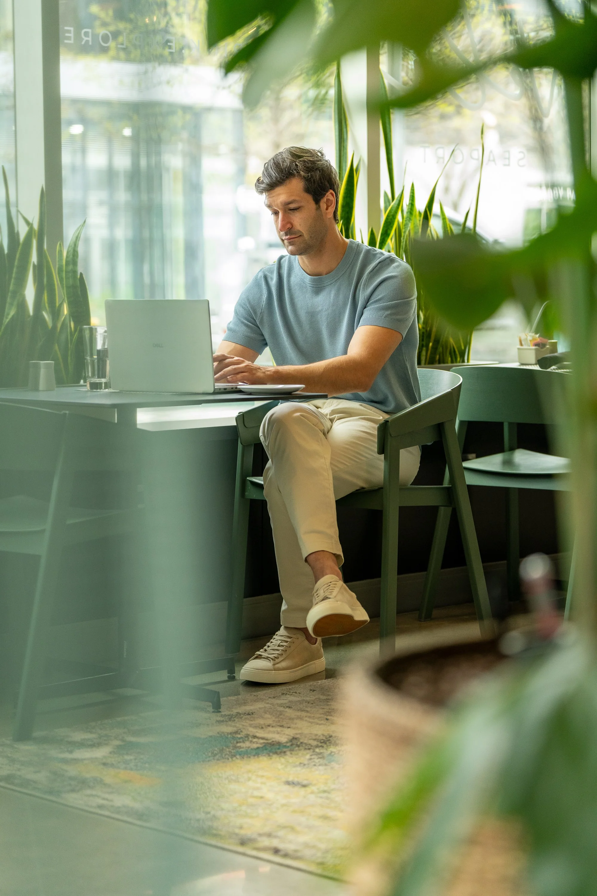 A man sitting at a table in a cafe working on a laptop, seen through a window with plants partially obscuring the view.
