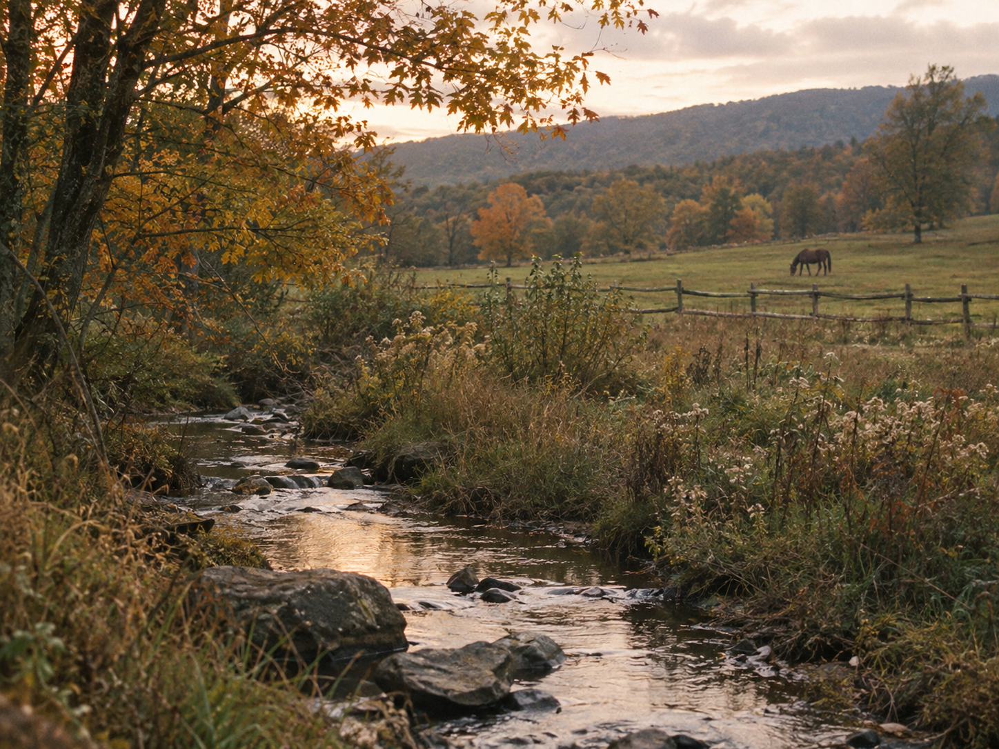 A peaceful landscape featuring a small stream flowing through a grassy field with trees showing fall foliage, a wooden fence, and a horse grazing in the distance under a partly cloudy sky.