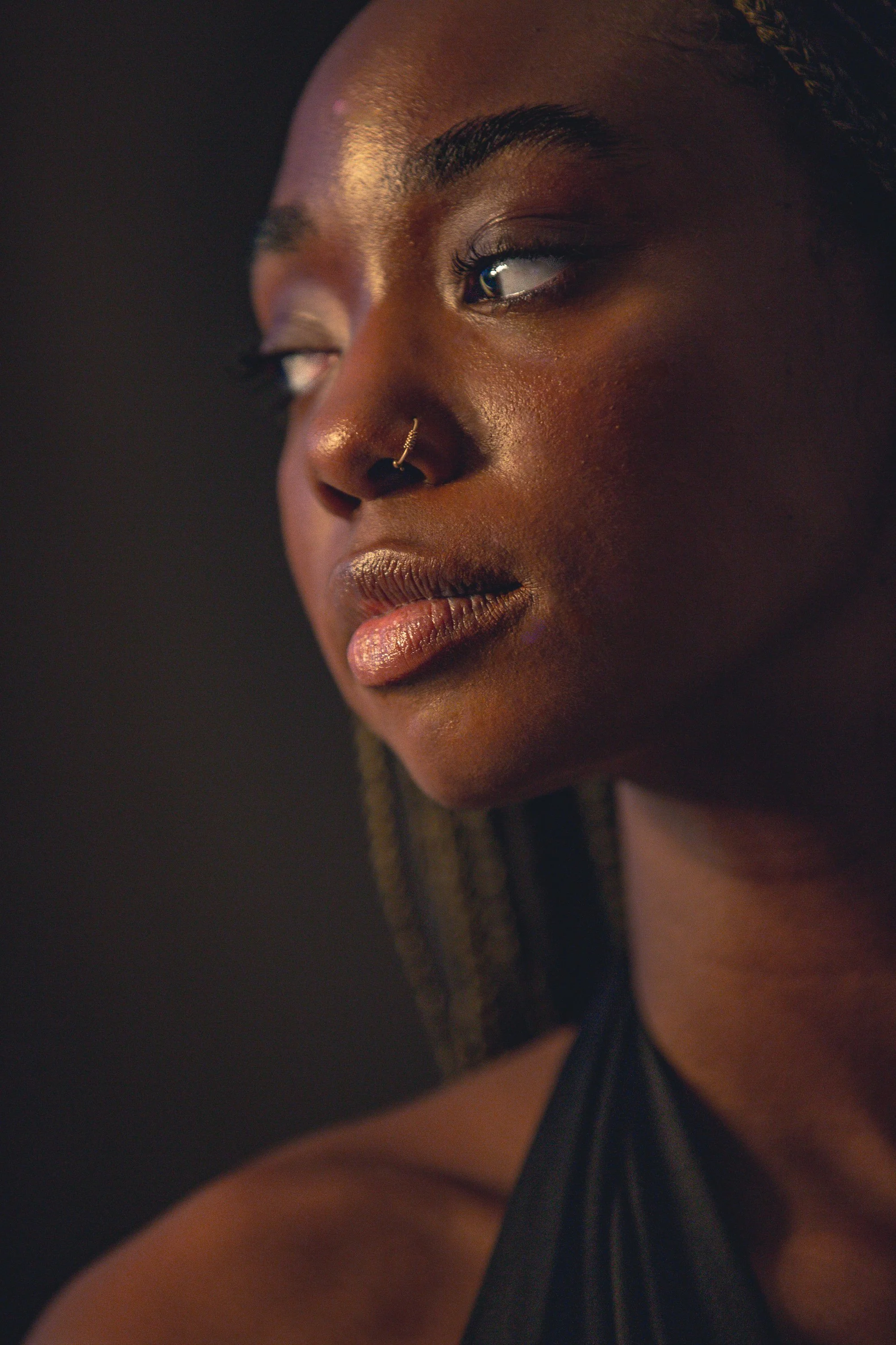 Close-up of a woman with dark skin, wearing a nose ring, with makeup, looking to the side, with braided hair, and a dark background.