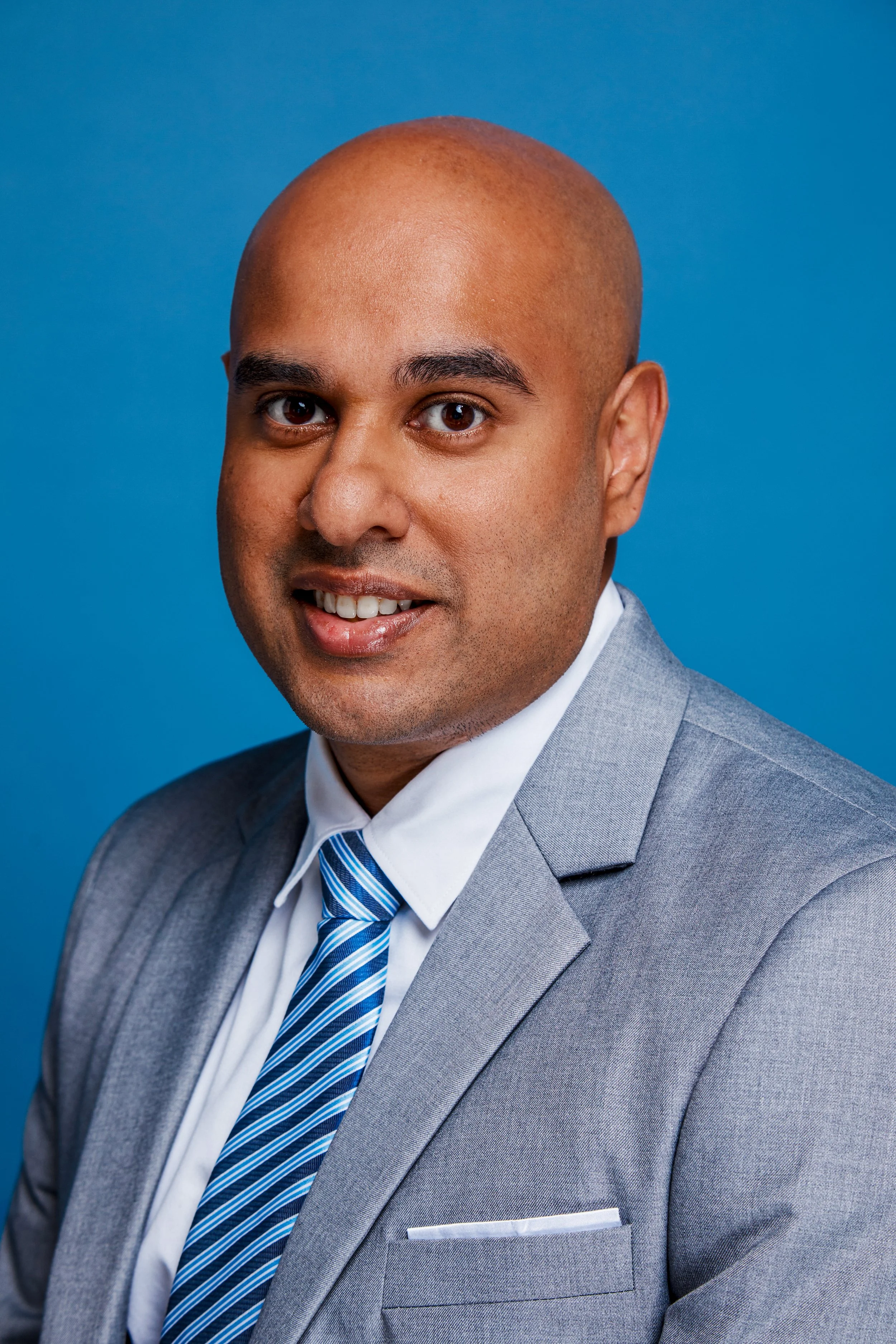 Headshot of a man with a shaved head wearing a gray suit, white shirt, and a blue striped tie, against a blue background.