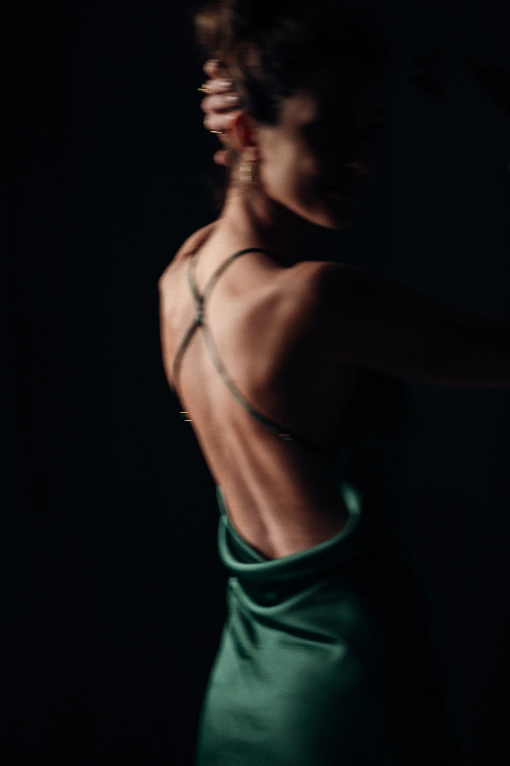 A woman with short hair and earrings, wearing a spaghetti strap top, standing in front of a dark background.
