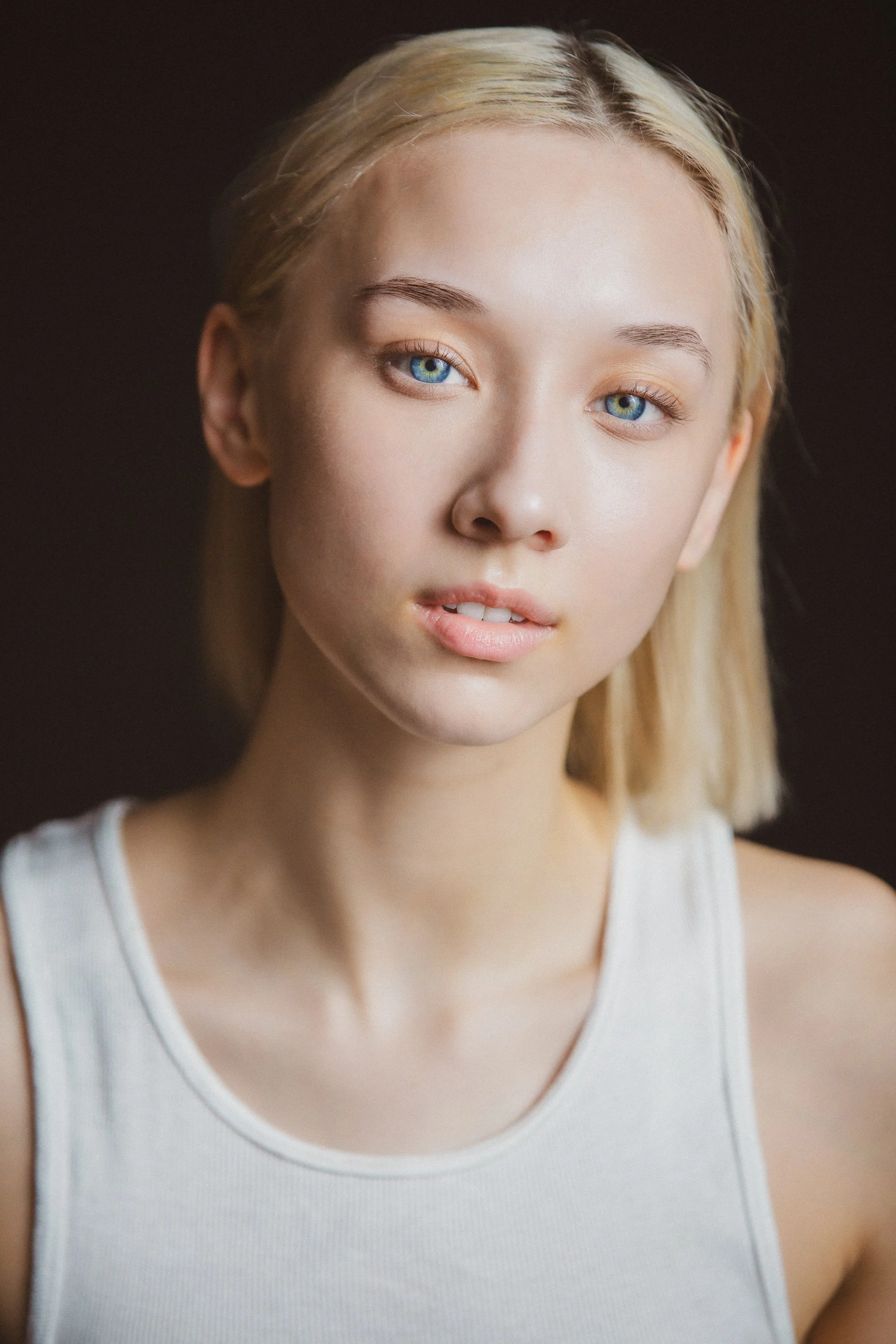 Close-up of a young woman with blue eyes, blonde hair, and light skin wearing a white sleeveless top.