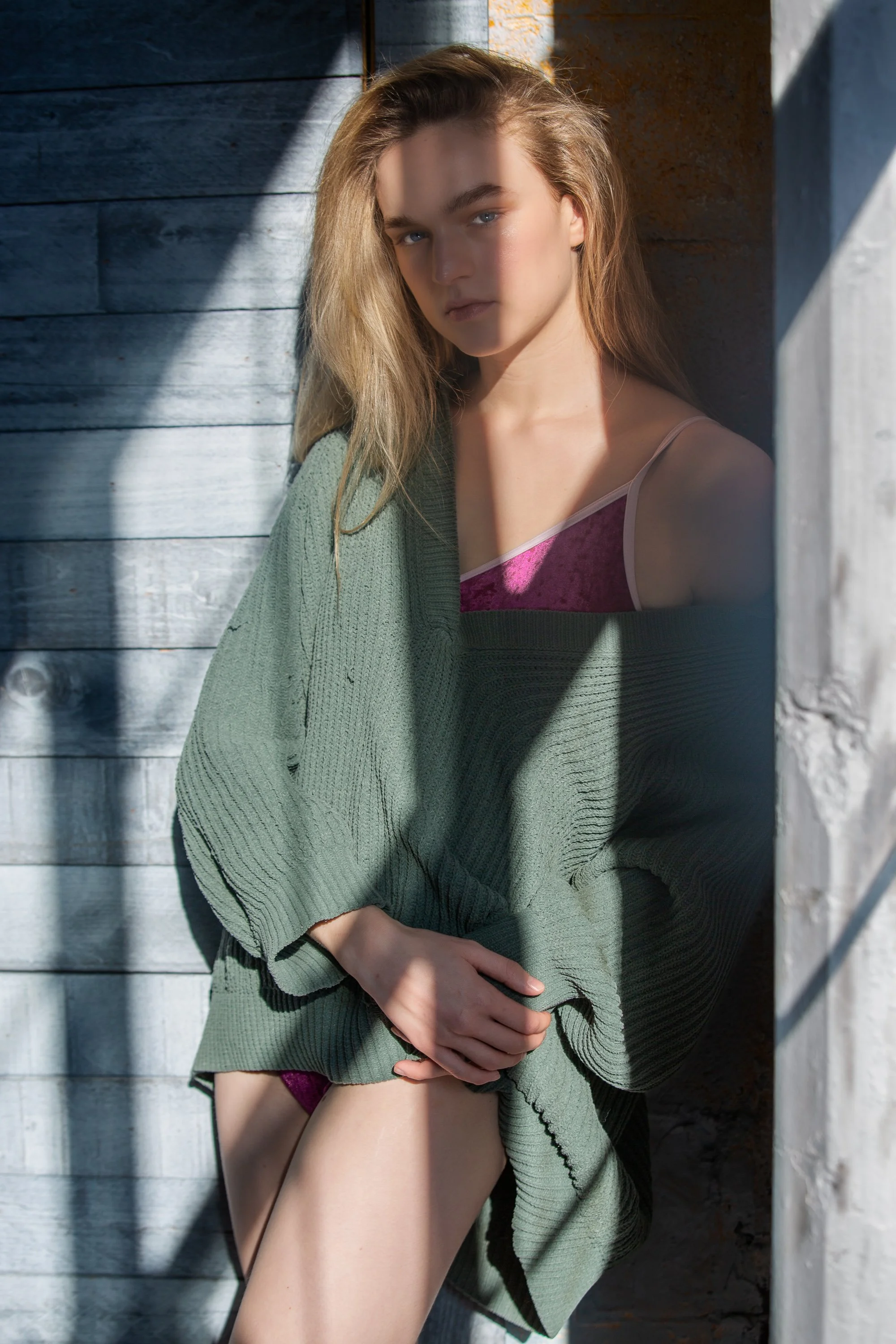 A woman leaning against a wooden wall, wearing a green sweater and pink pajamas, with sunlight casting shadows on her face and body.