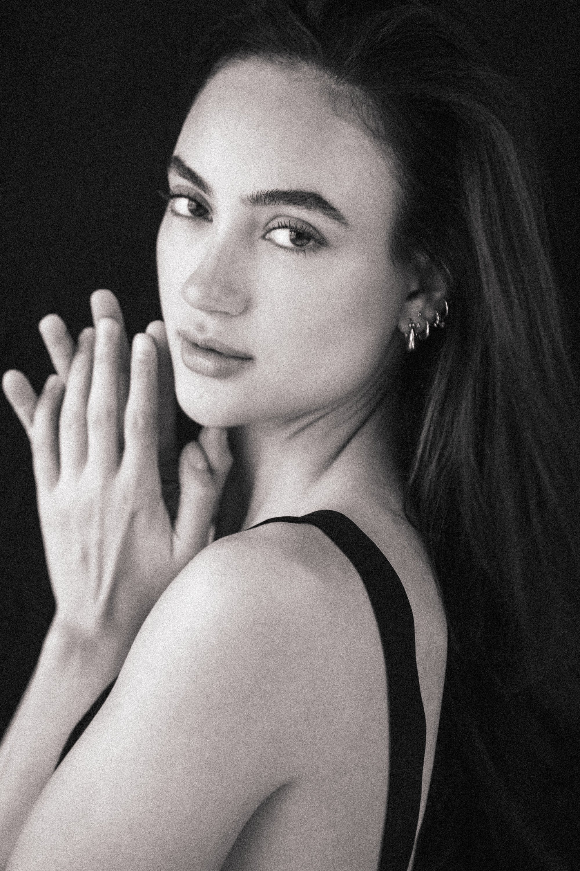 Black and white portrait of a young woman with long hair, multiple earrings, and wearing a sleeveless top, posing with her hands near her face on a dark background.