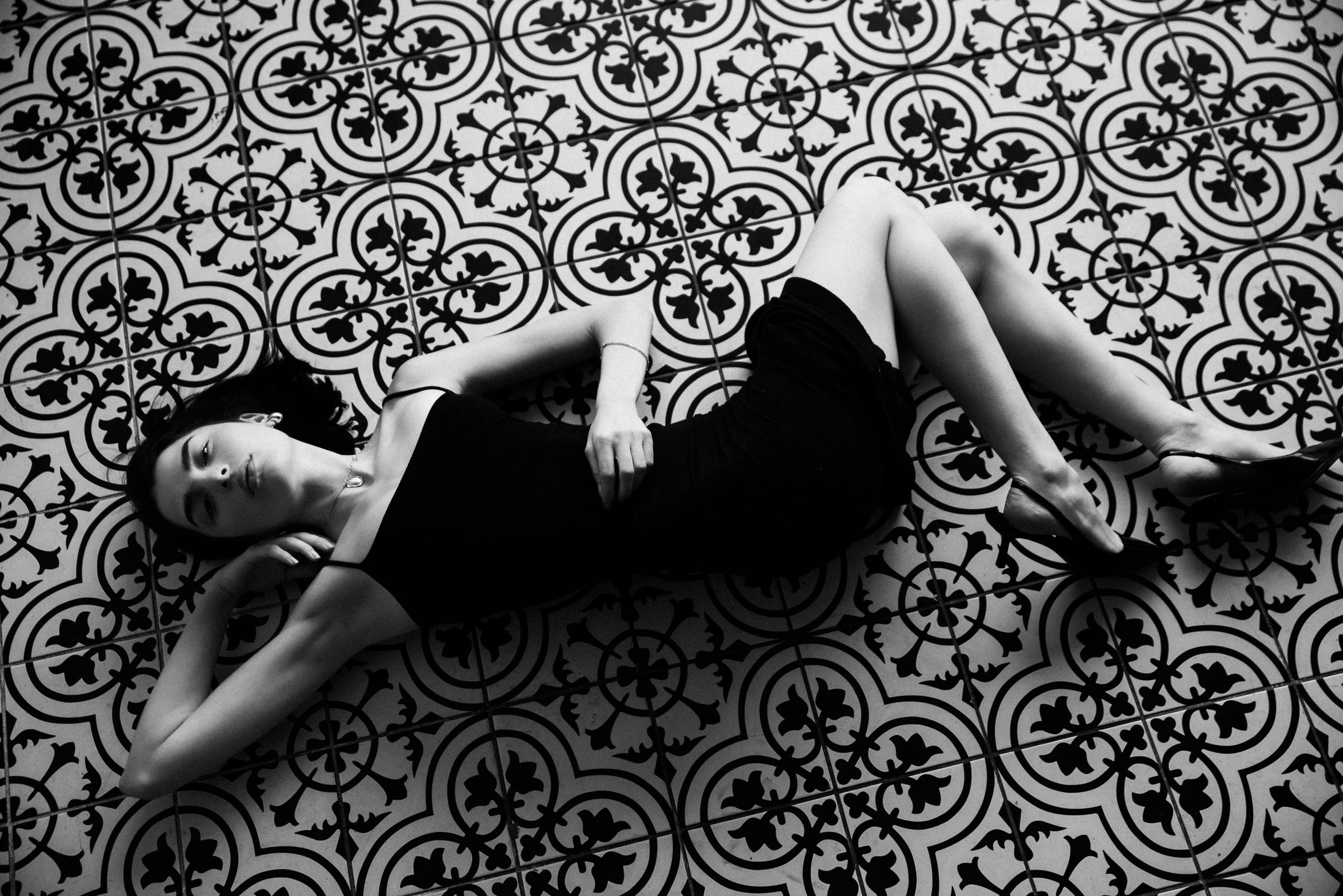 A woman lying on a patterned floor with her eyes closed, wearing a black dress and high heels.