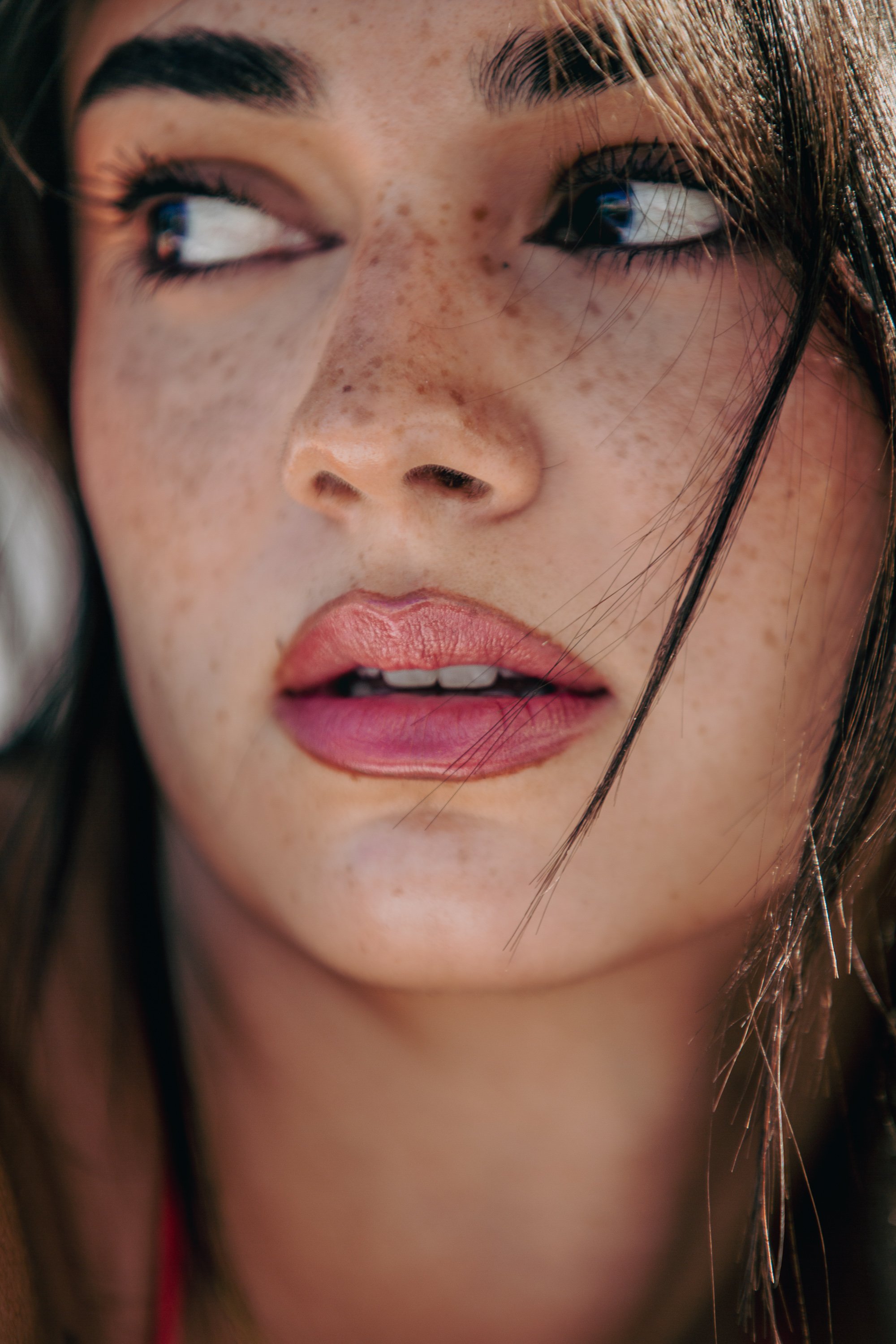A close-up photograph of a young woman with dark, straight hair partially covering her face. She has fair skin with freckles and is looking sideways, exposed to natural light.