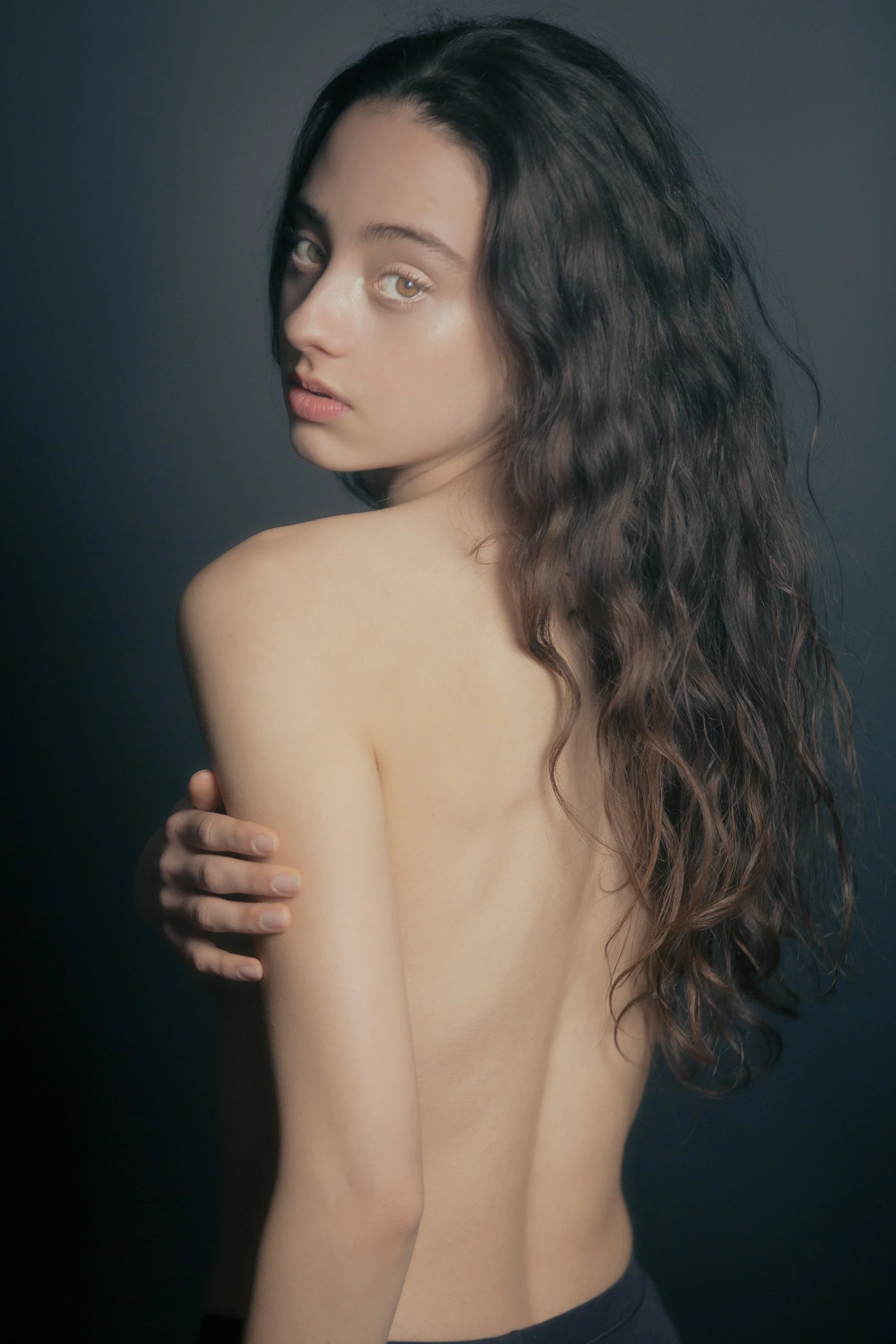 A woman with long wavy brown hair and fair skin is posing against a dark background, covering her chest with her arm and looking at the camera. The lighting highlights her facial features and shoulder.