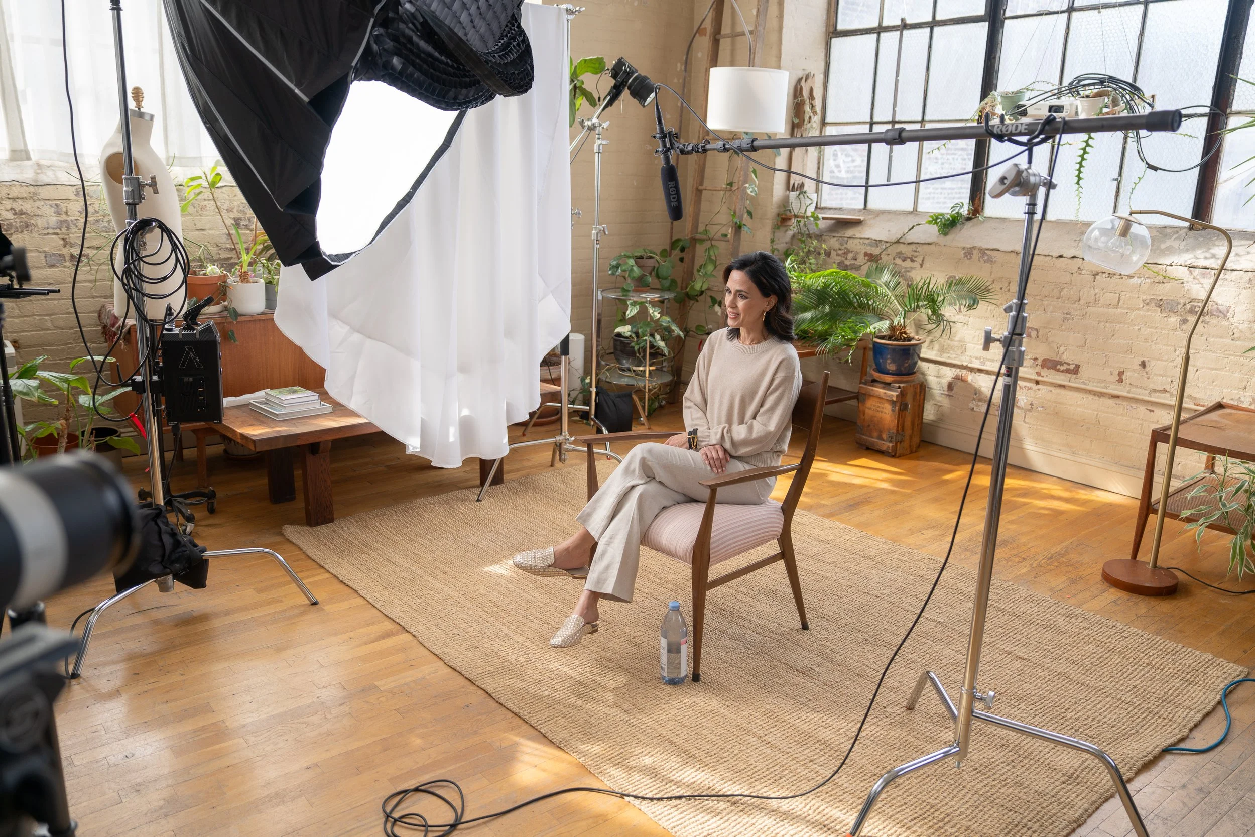 A woman sitting on a chair in a photography studio surrounded by filming equipment, plants, and natural light coming through large windows.