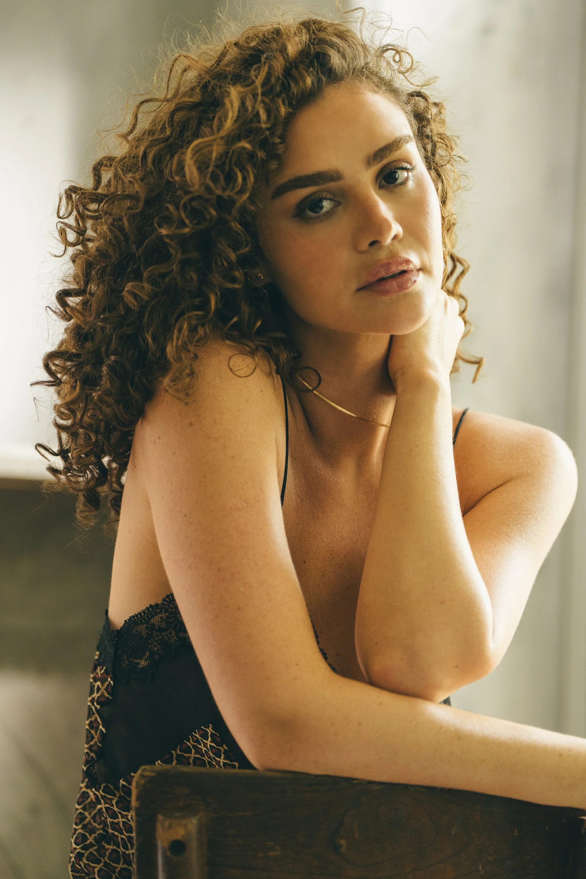 A woman with curly hair and freckles, wearing a black lace top, sitting with her hand on her neck, looking at the camera in a softly lit setting.