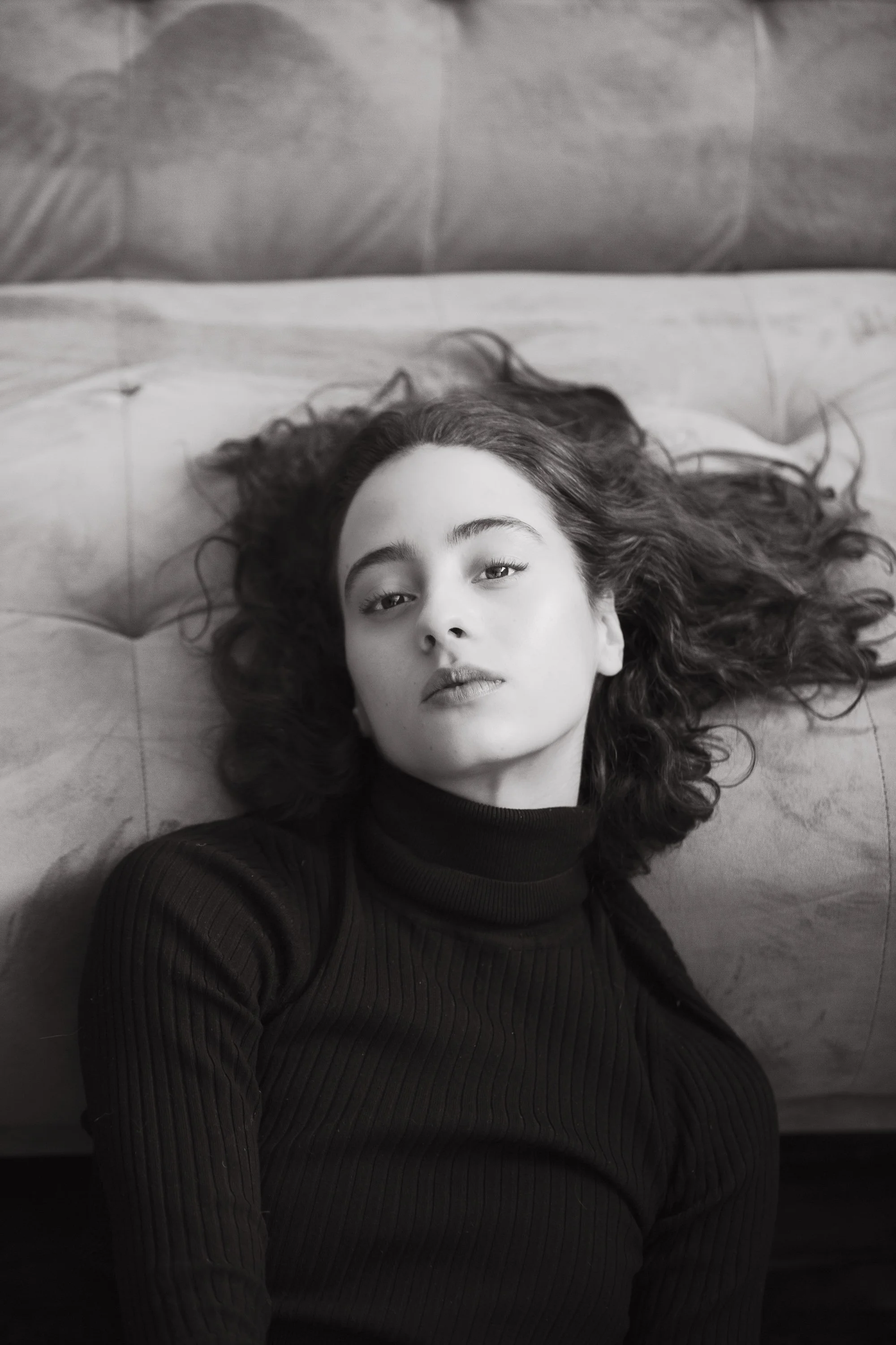 A young woman with wavy hair lying on a bed, looking directly at the camera.