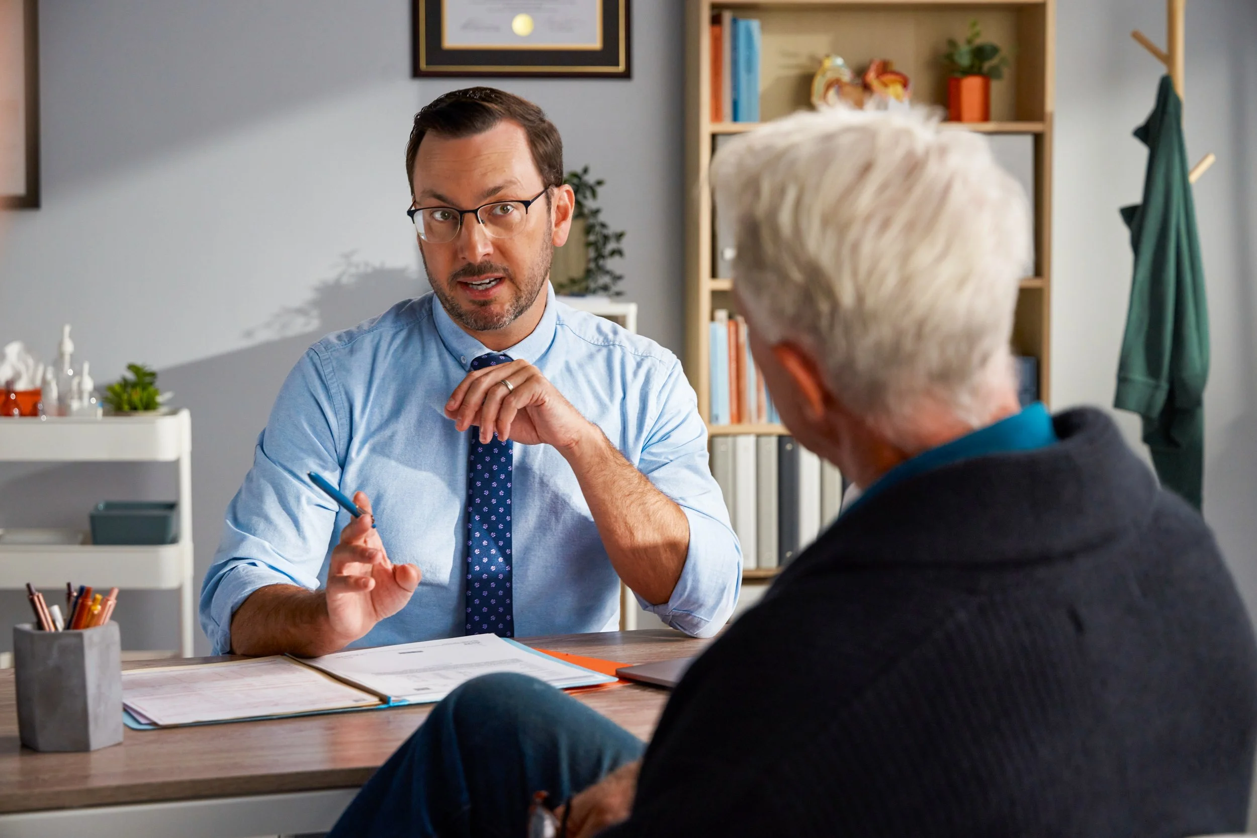 A middle-aged man with glasses and a beard wearing a light blue shirt and a polka dot tie is sitting at a desk, speaking to an older man with white hair, seen from the back. The setting appears to be an office or consulting room with bookshelves and framed artwork in the background.
