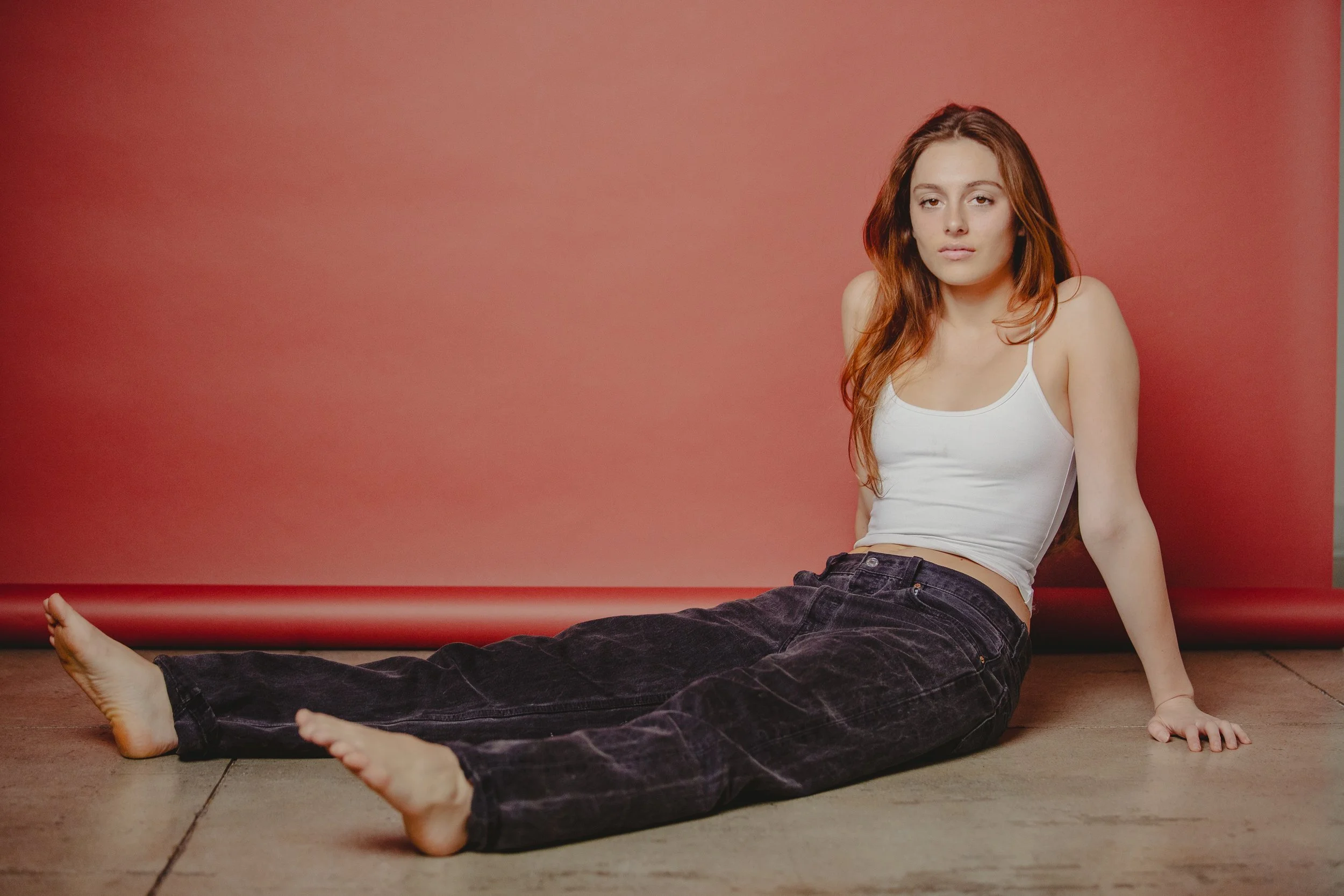A woman with long red hair sitting on the floor against a red wall, wearing a white tank top and black pants, with her legs extended and hands resting on the ground.
