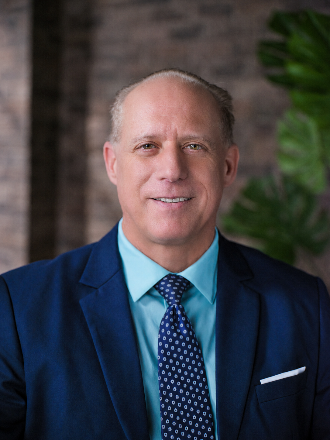 A middle-aged man with light skin, short bald hair, wearing a navy suit with a light blue shirt and a dark blue tie with small white dots, smiling in front of a blurred background with wooden panels and green foliage.