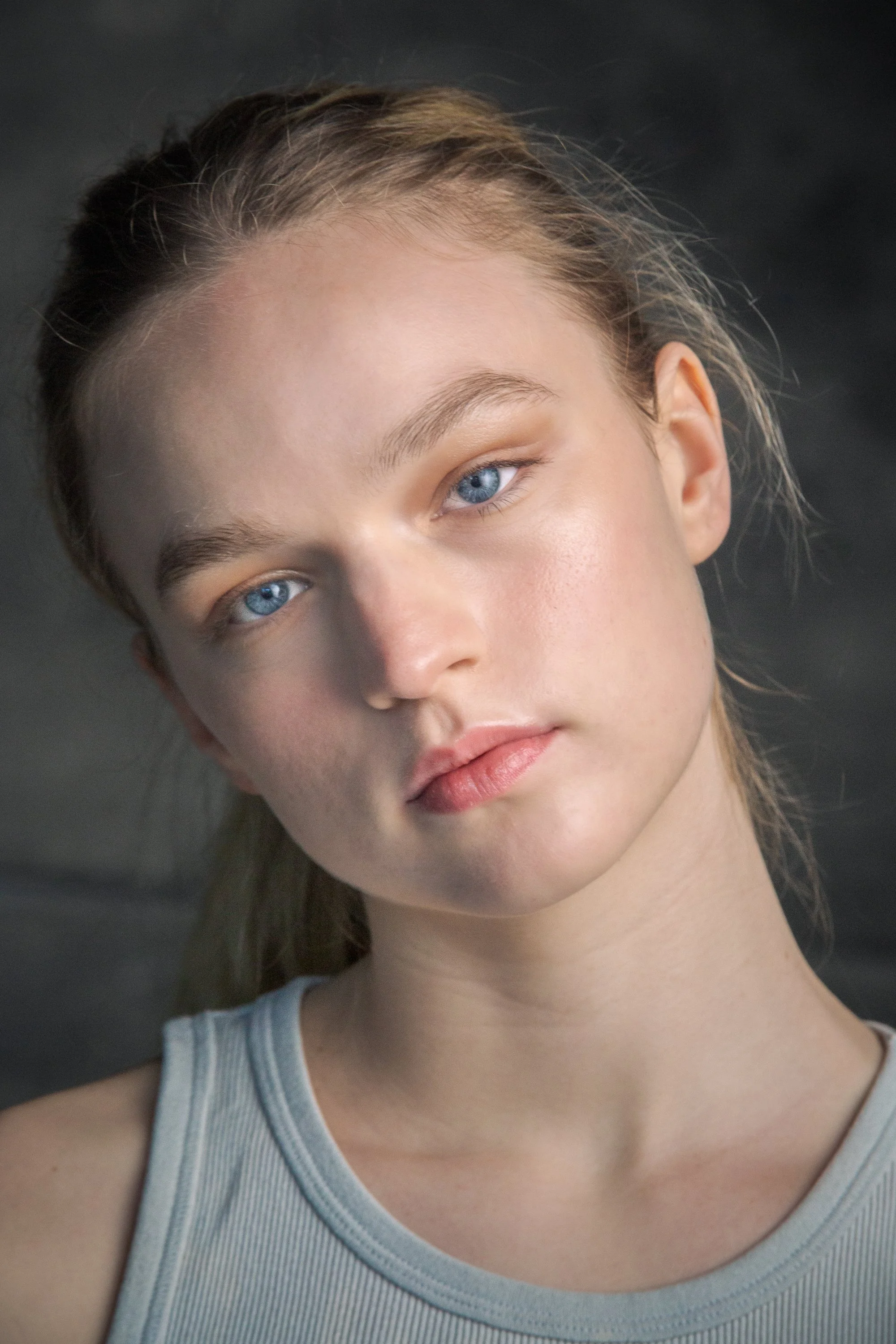 Close-up of a young woman with light skin, blue eyes, and long hair, wearing a gray sleeveless top, against a dark background.