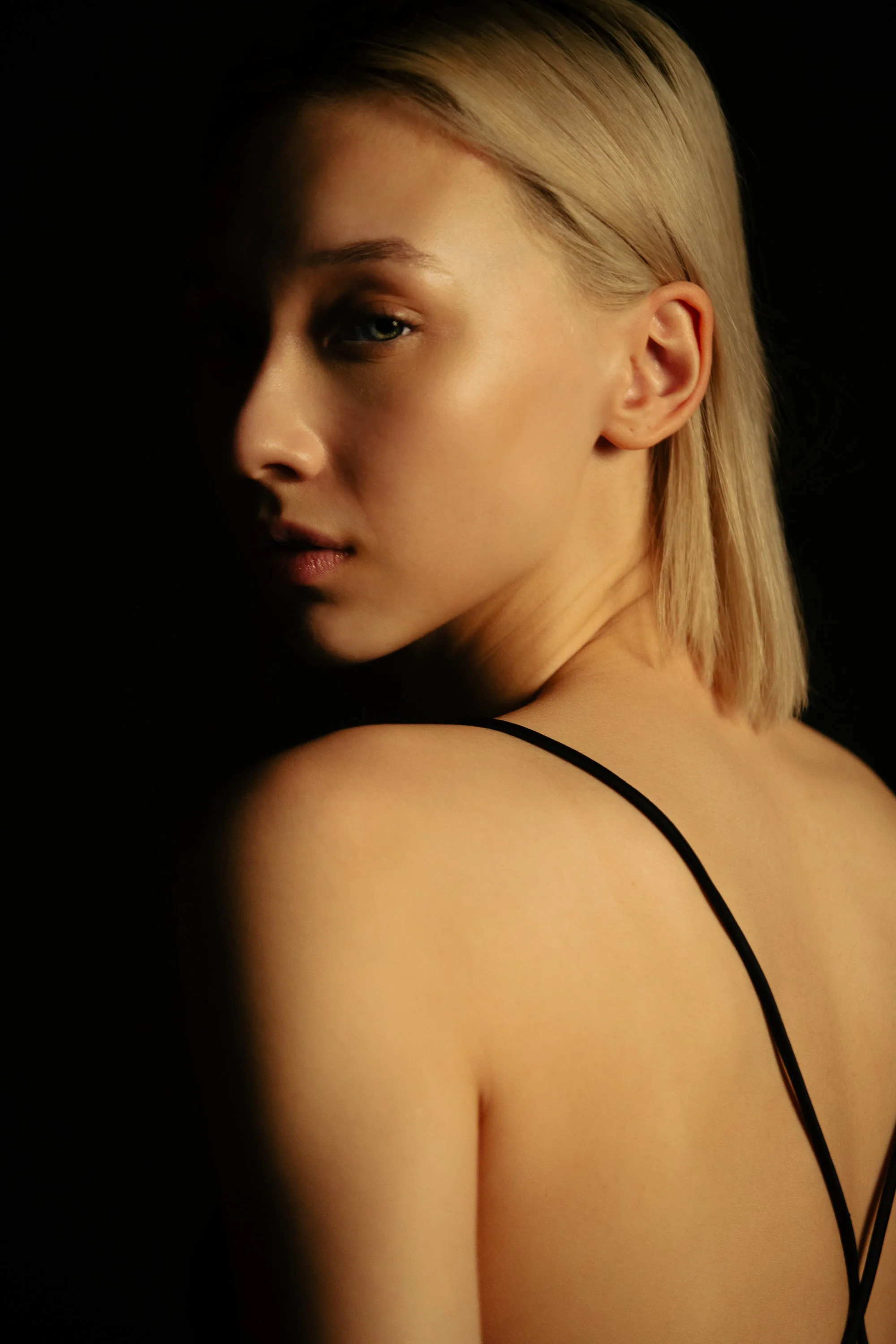 A close-up portrait of a woman with blonde hair, partially shadowed on the left side of her face, wearing a black strap top, against a dark background.