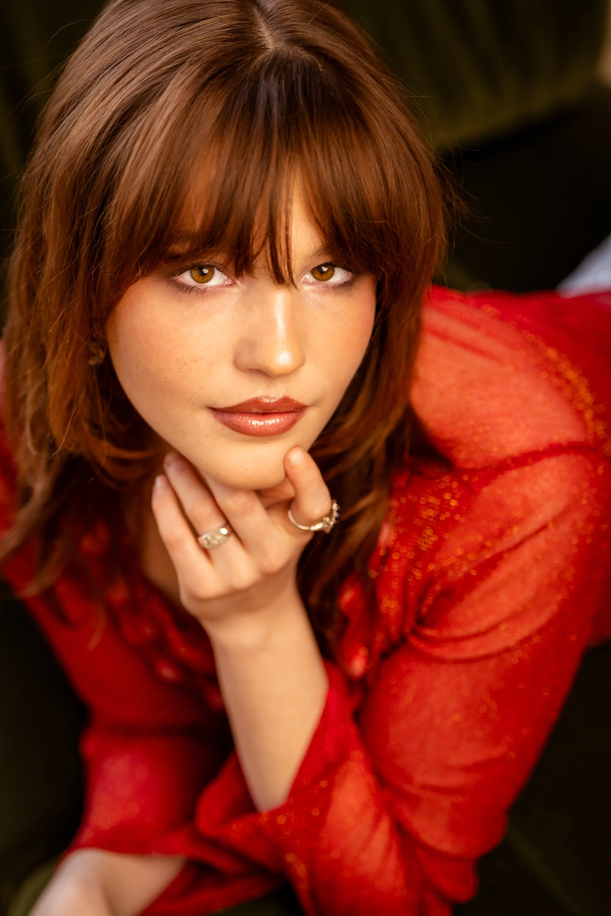 A woman with light brown hair, brown eyes, and wearing a red, sparkly dress, poses with her hand near her chin and gazes into the camera.
