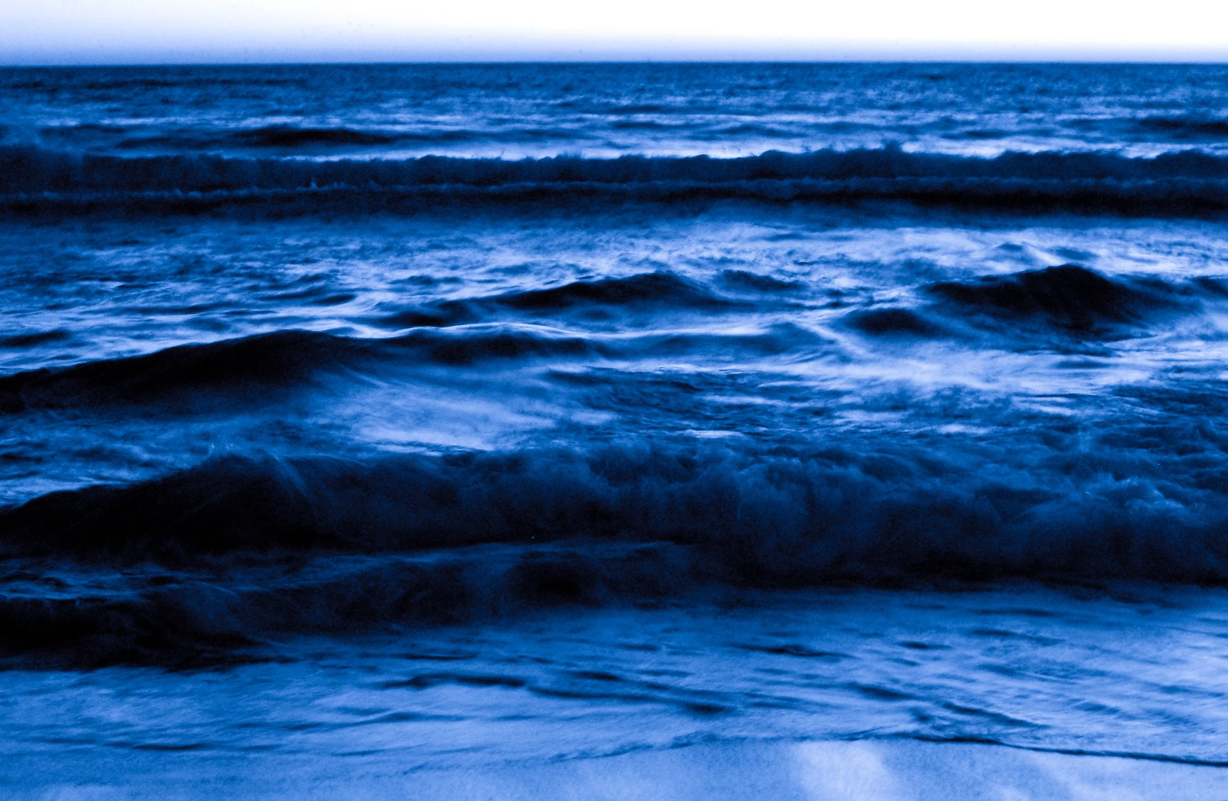 Dark ocean waves with a calm horizon in the background, under a clear sky.
