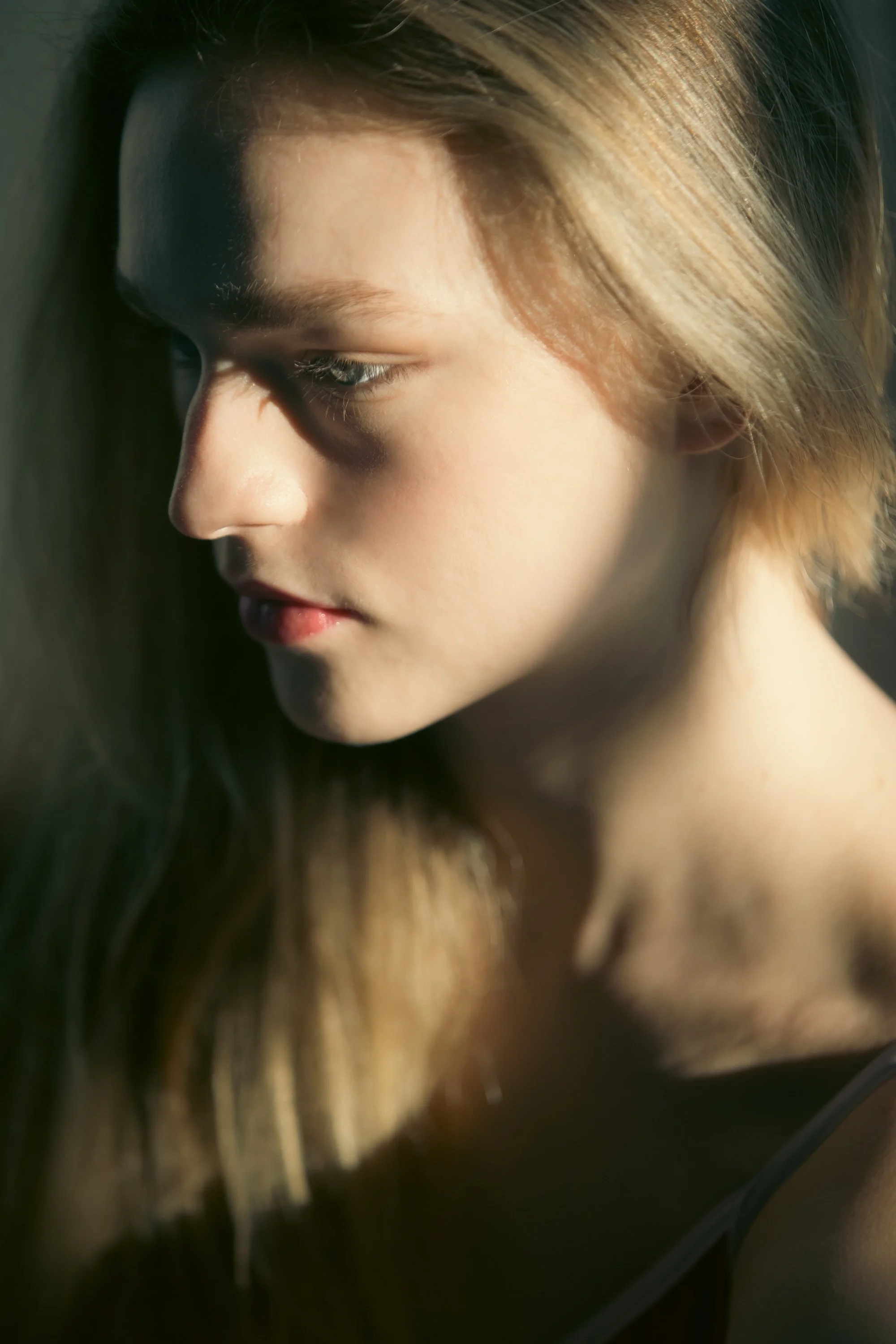 Close-up of a young woman's face in profile, with long hair falling over her shoulder, looking downward with sunlight illuminating her hair and skin.