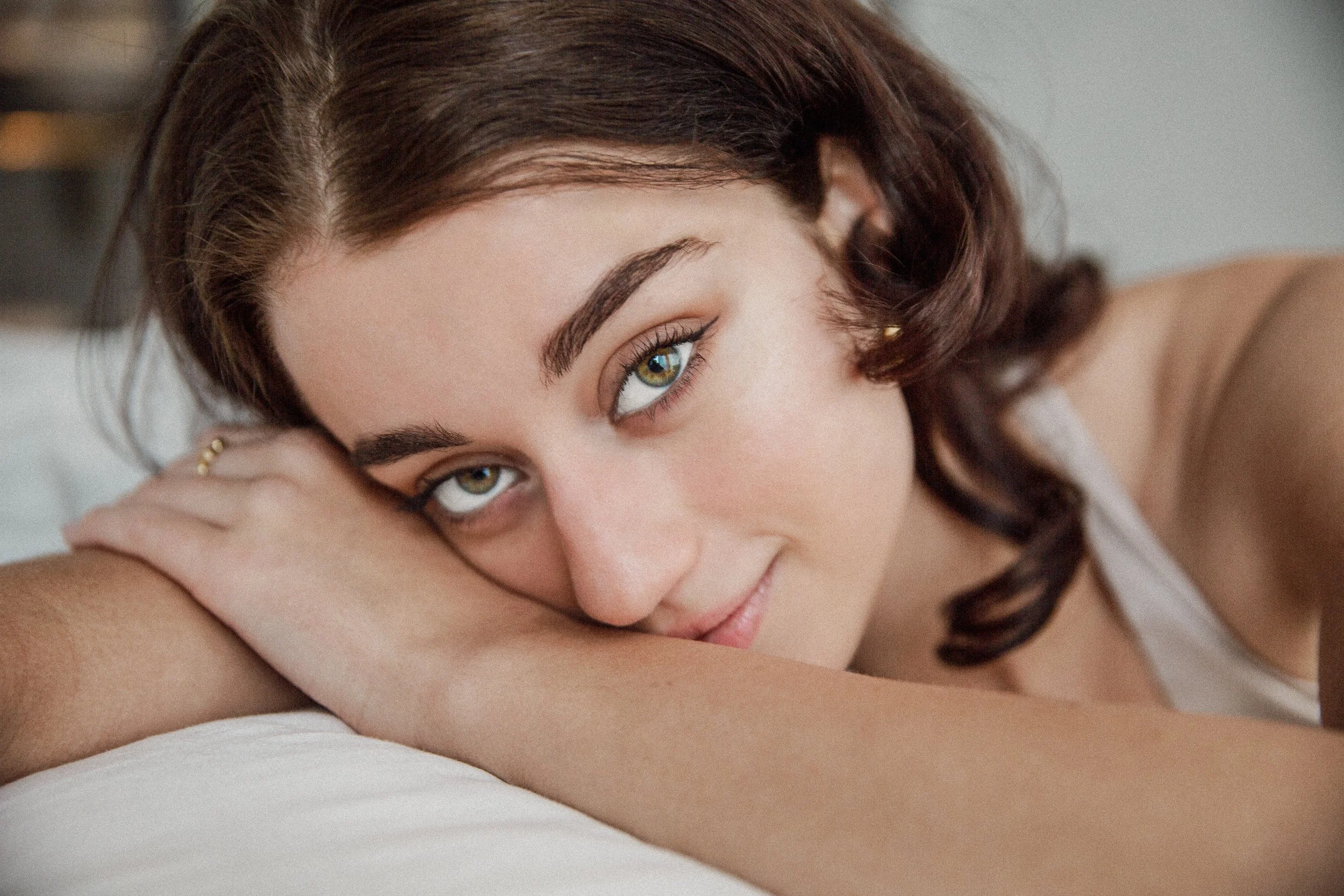 A close-up of a woman with brown hair and green eyes resting her head on her crossed arms, looking into the camera.