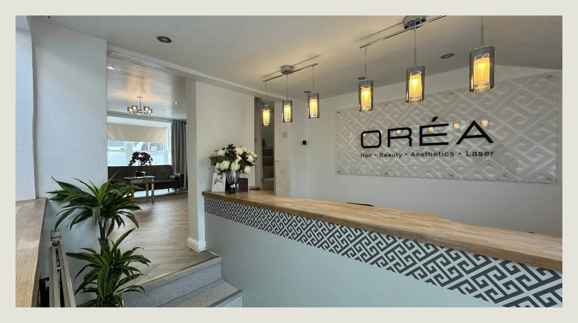 Interior of a salon named OREA, with a reception desk, hanging pendant lights, a floral arrangement, lobby seating area, and a patterned wall display with the salon's name and services listed.