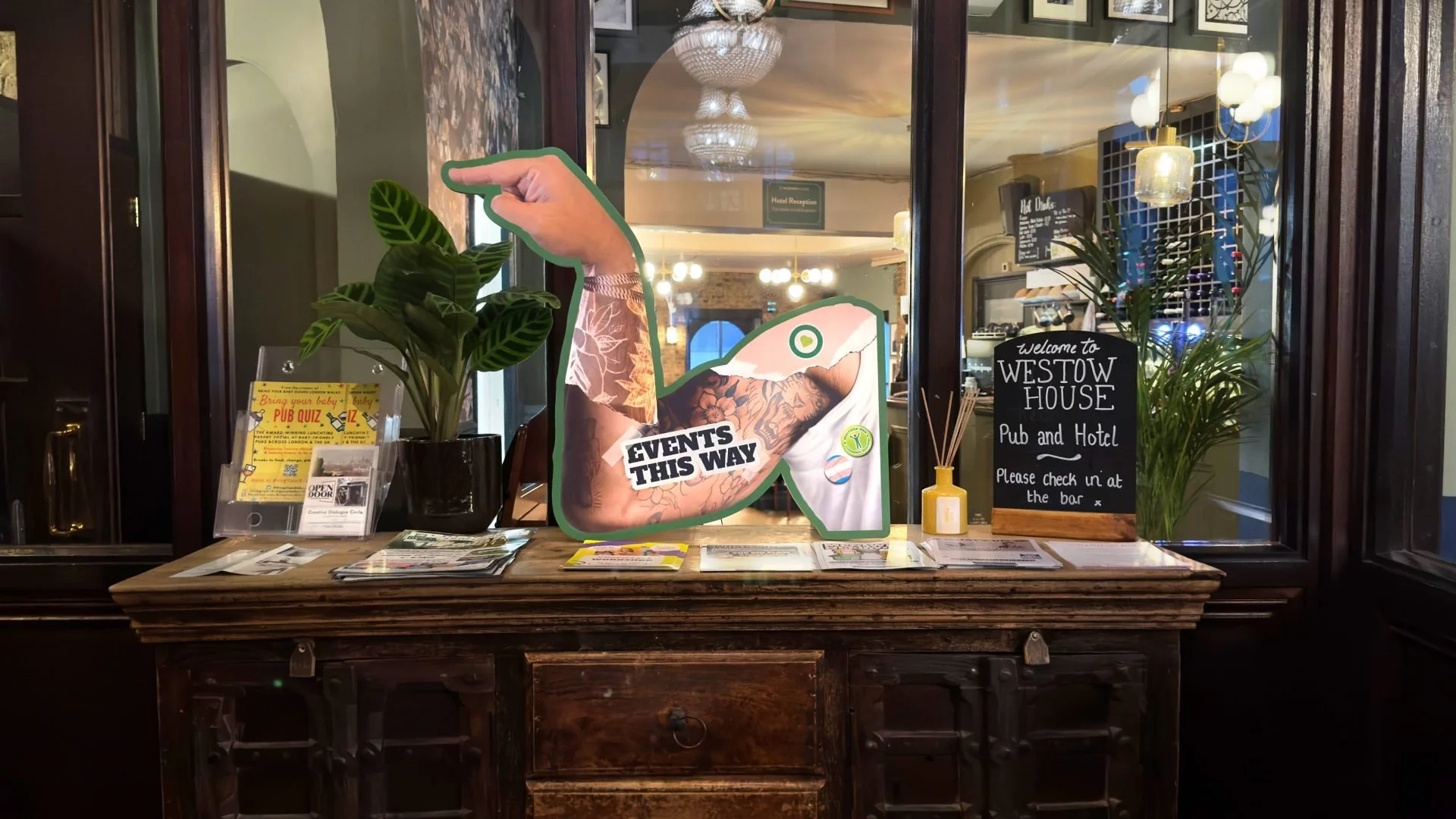 The image shows a wooden reception desk with plants, brochures, and a sign that says "Welcome to Westow House Pub and Hotel." Behind the desk, there is a large window with reflections of chandeliers and hanging lights inside the building, and signs a