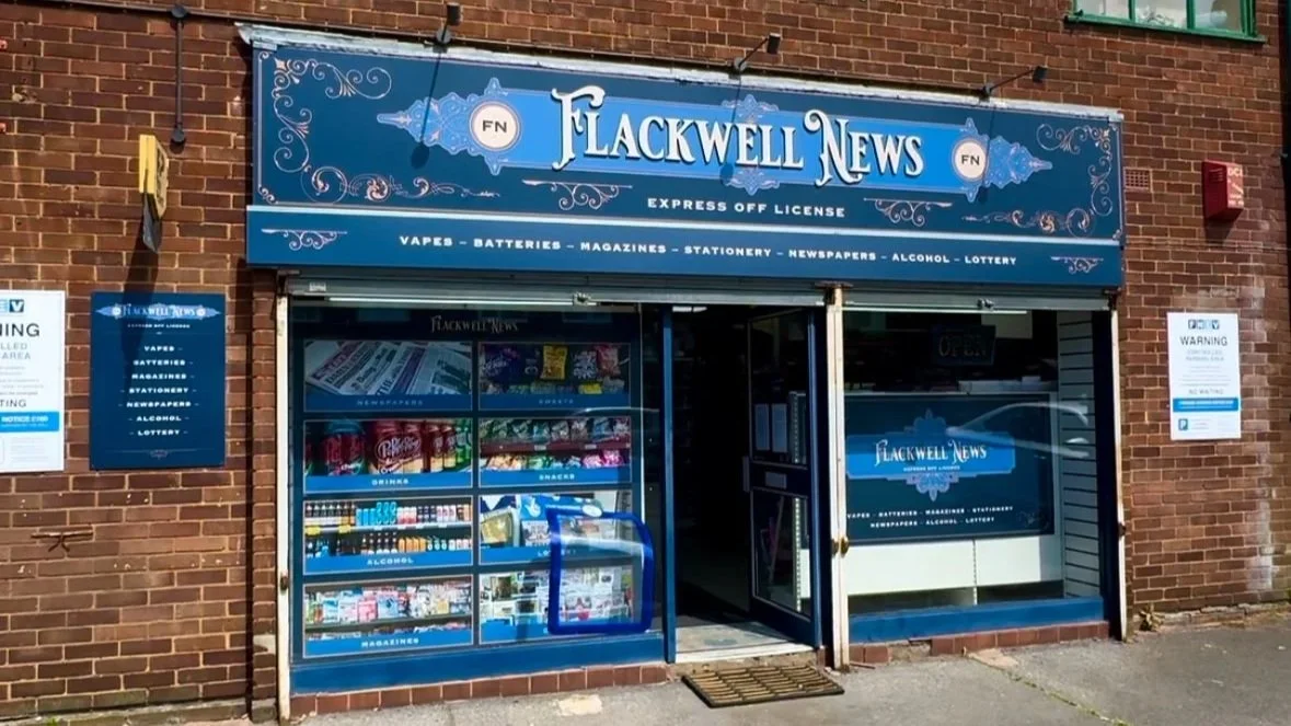 Front view of a convenience store called 'Flackwell News' in a brick building, selling newspapers, magazines, snacks, drinks, and tobacco products.