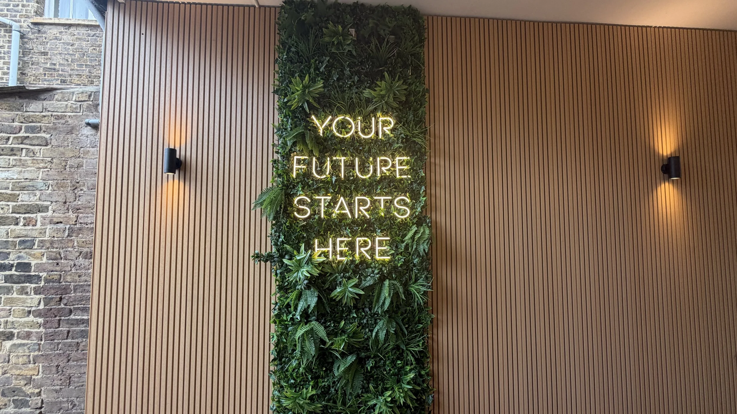 Warm-white illuminated motivational wall sign surrounded by green foliage and timber slat panelling.JPG