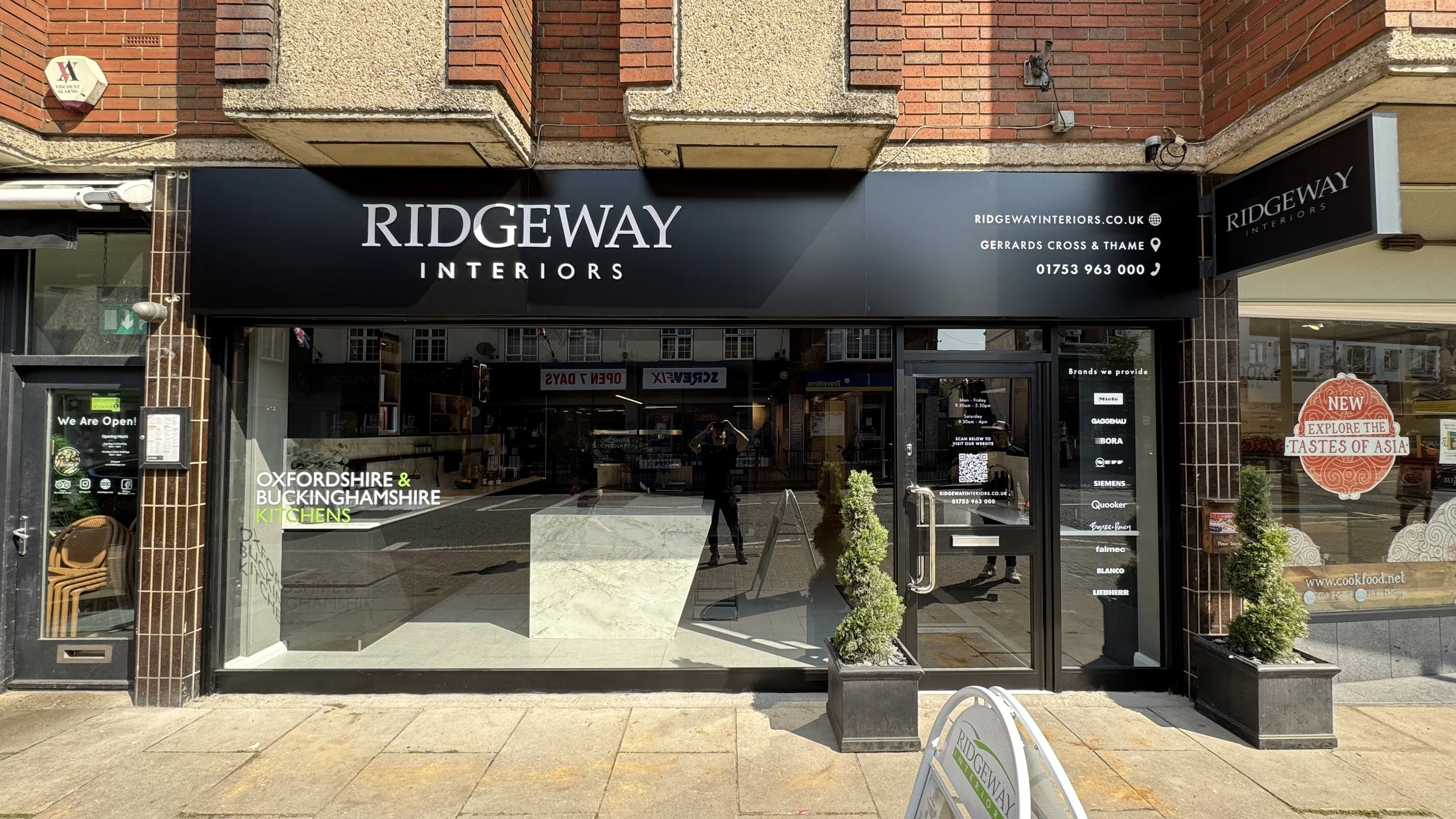  storefront with a black sign displaying the name 'Ridgeway Interiors' in white letters, surrounded by brick and concrete. There are small windows and a glass door with signage displaying brands and opening hours. Plants are placed outside, and the r