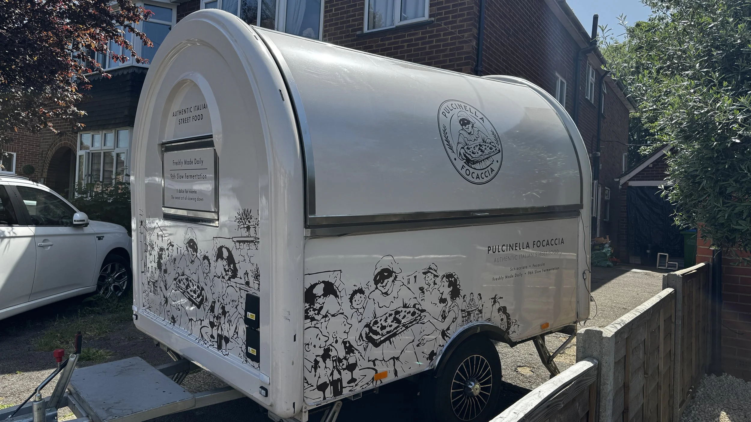A white food trailer with black line illustrations and the logo 'Pulcinella Focaccia,' parked on a street in front of residential buildings and trees.