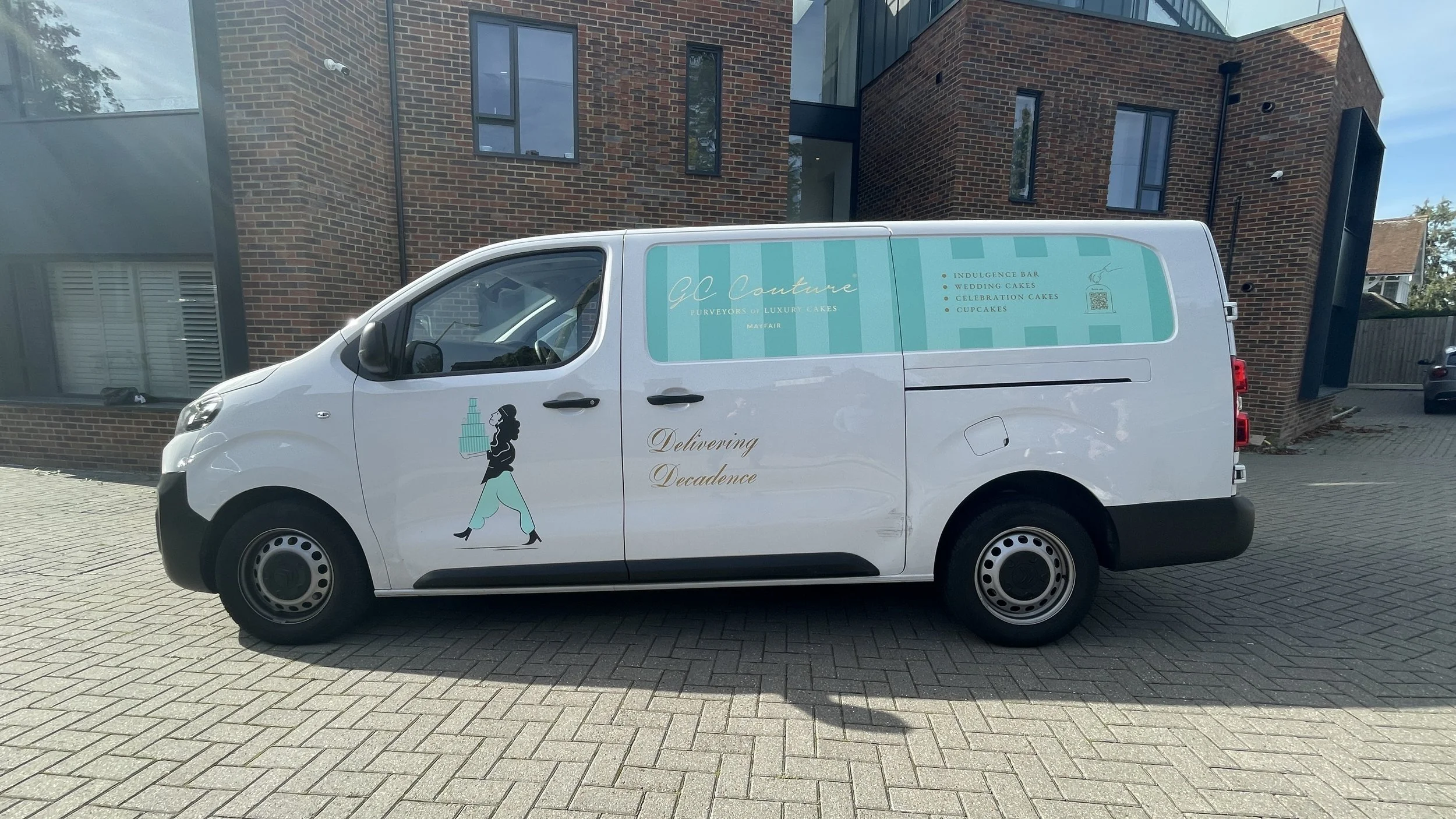 White delivery van parked on brick-paved street in front of a modern brick building, with branding for GC Couture Purveyors of Luxury Cakes, featuring a stylized silhouette of a woman carrying a tiered cake, and the text "Delivering Decadence" on the
