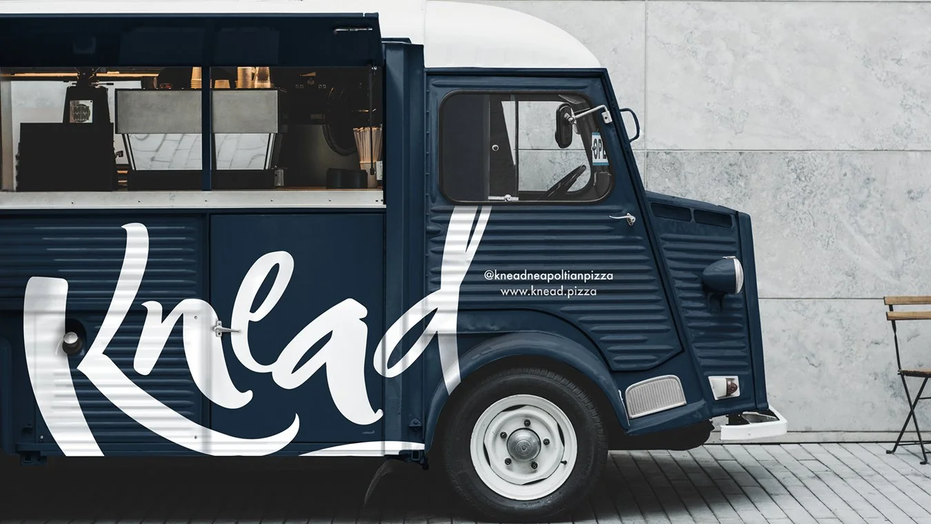 A blue food truck with white cursive text and a menu for a pizza business.