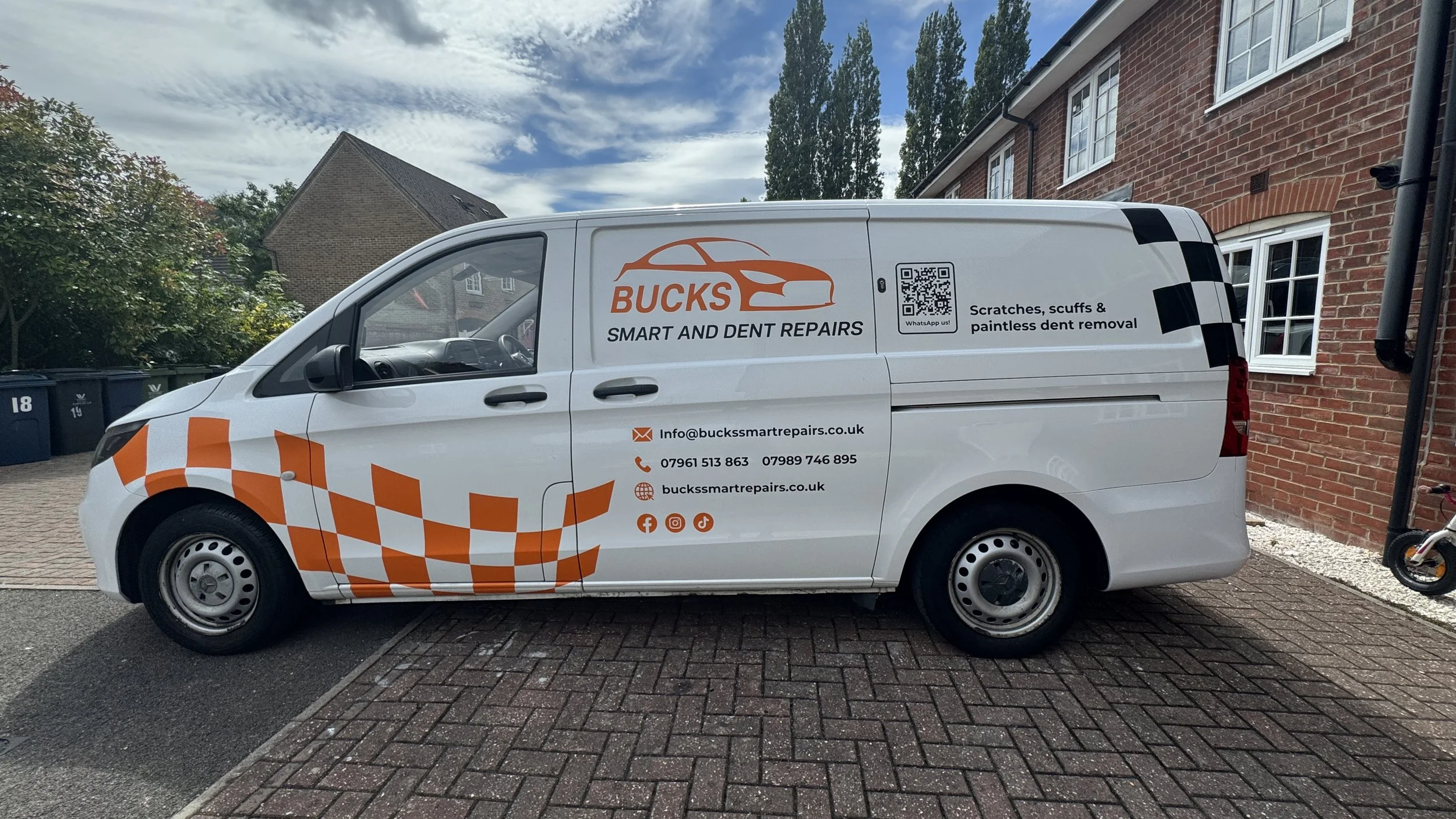 White delivery van parked outside a brick house with a red brick wall. The van has orange and black checkered patterns and company branding for Bucks Smart and Dent Repairs, including contact information, social media icons, and a QR code. The house 