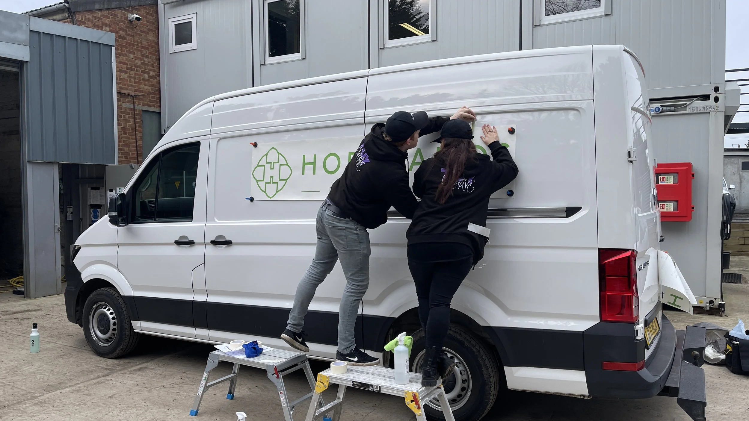 Two people are working on wrapping a white van with a green logo and text. They are pinning a large decal onto the side of the vehicle. The scene is outdoors in a lot with some tools and supplies nearby, and a building with windows in the background.