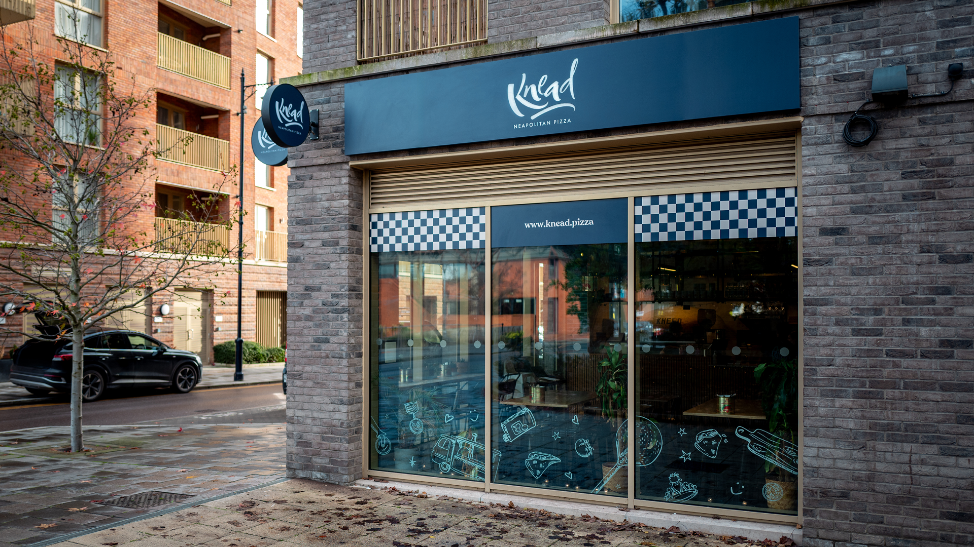 Exterior of a Neapolitan pizza restaurant named Knead with a dark blue sign and logo, large glass windows decorated with food illustrations, and brick building on a city street.
