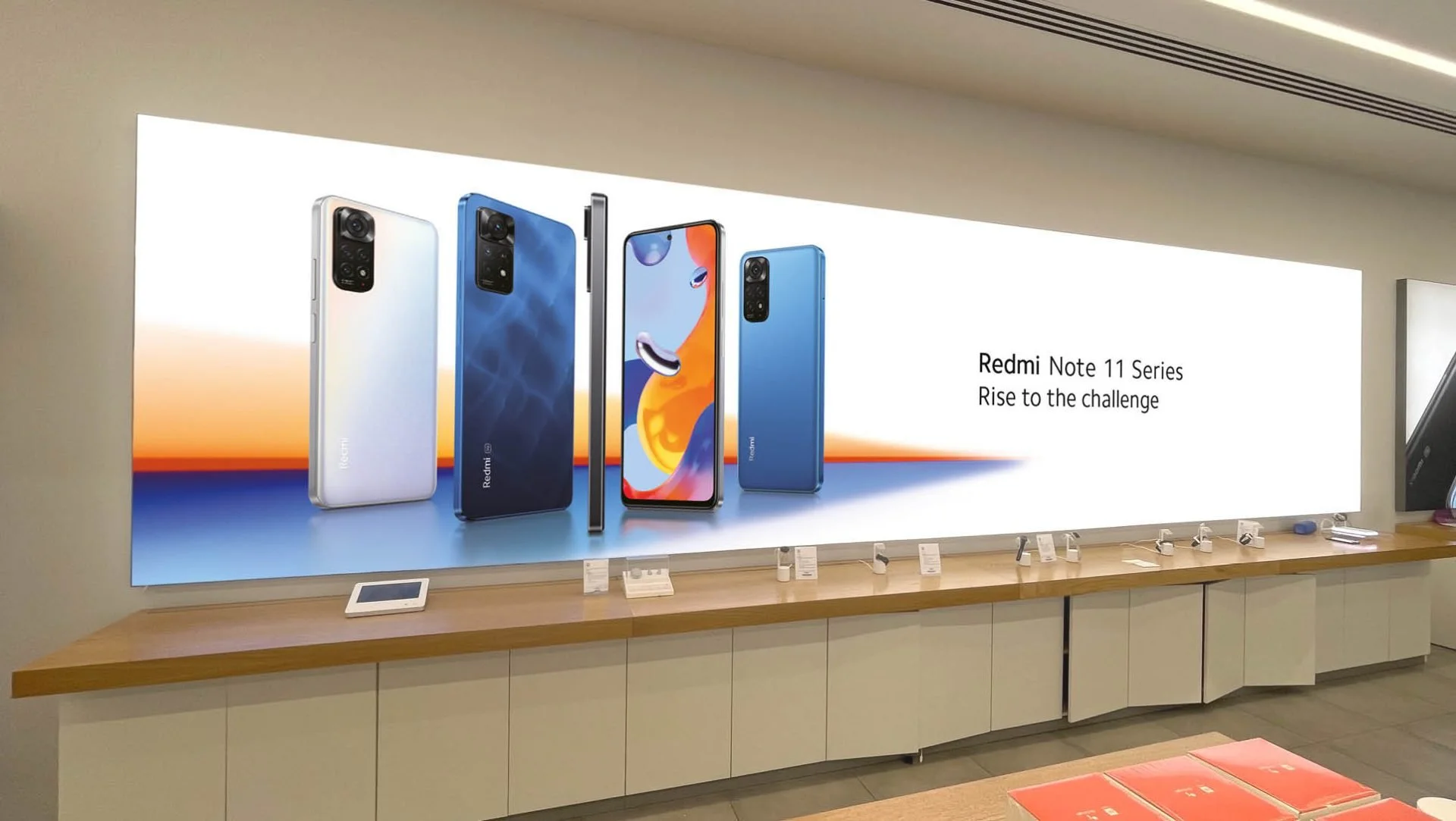 A digital display in a store shows various models of the Redmi Note 11 Series smartphones. The ad features four phones in different colors, with the slogan 'Rise to the challenge'.