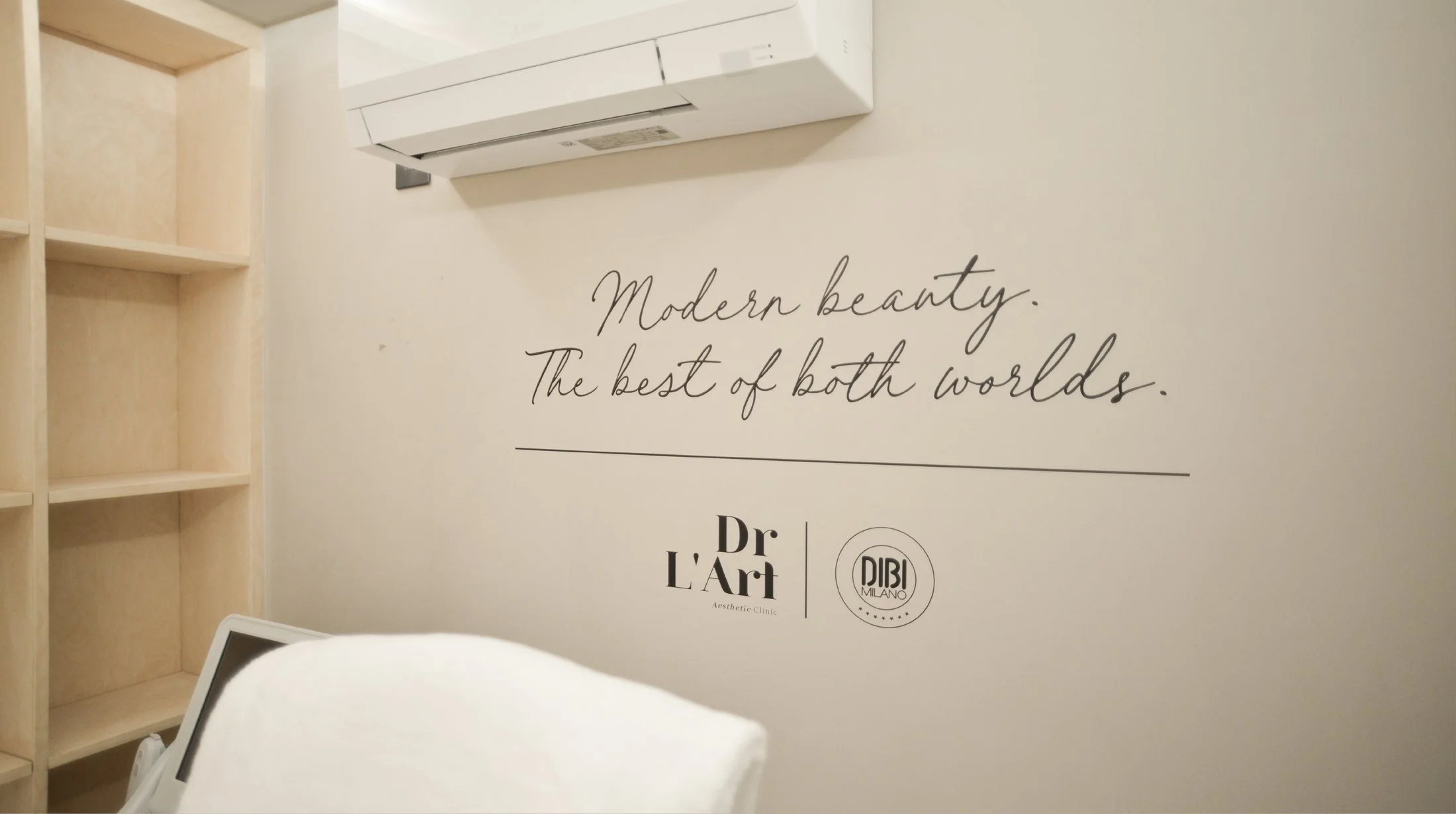 Interior of a clinic room with a wall decal that says 'Modern beauty. The best of both worlds.' and logos for Dr. L'Art and DIBI Milano. Part of a computer screen and a person’s arm are visible in the foreground.
