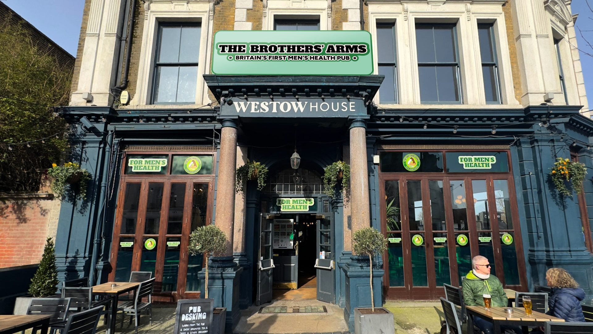 The exterior of a pub called Westow House, featuring a sign for The Brothers' Arms, Britain's first men's health pub. The pub has large windows with green signs that read 'For Men's Health' and outdoor seating with tables and chairs. A man wearing a green jacket and sunglasses is seated in the outdoor area.