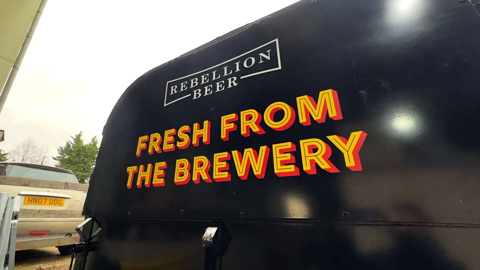 Black trailer with a sign displaying the text 'Rebellion Beer' and 'Fresh from the Brewery' in yellow and red lettering. Part of a parking lot with cars visible in the background.