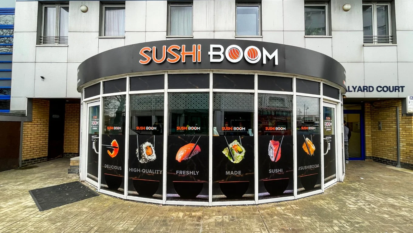 storefront with a black and white sign reading 'SUSHI BOOM' and large posters of sushi dishes on the windows.