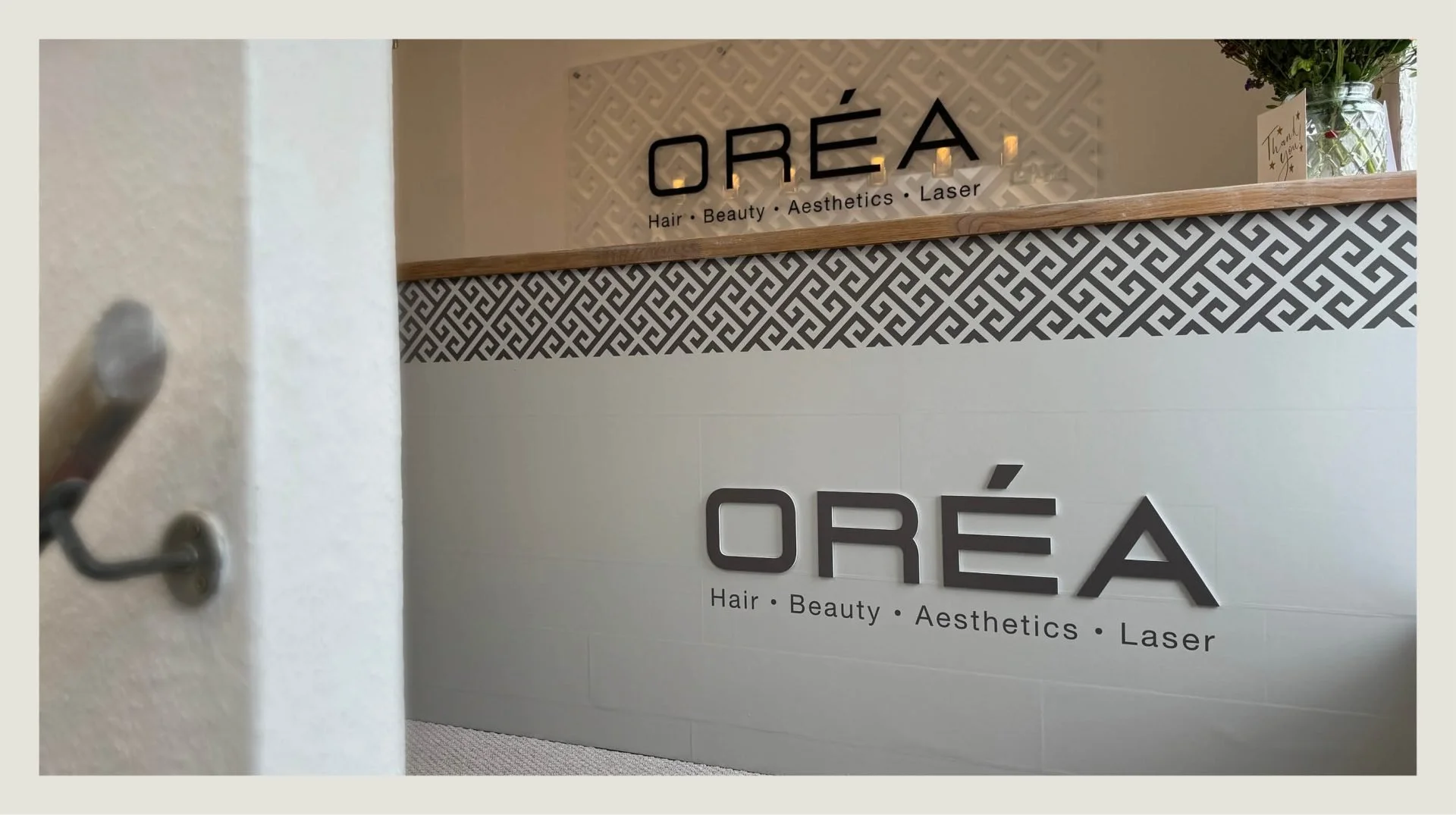 Sign for a beauty salon named 'Oréa' with services listed as Hair, Beauty, Aesthetics, and Laser. The sign is mounted on a wall with decorative black and white patterned tiles, and a wooden ledge is above it. A vase with flowers and a card are visibl