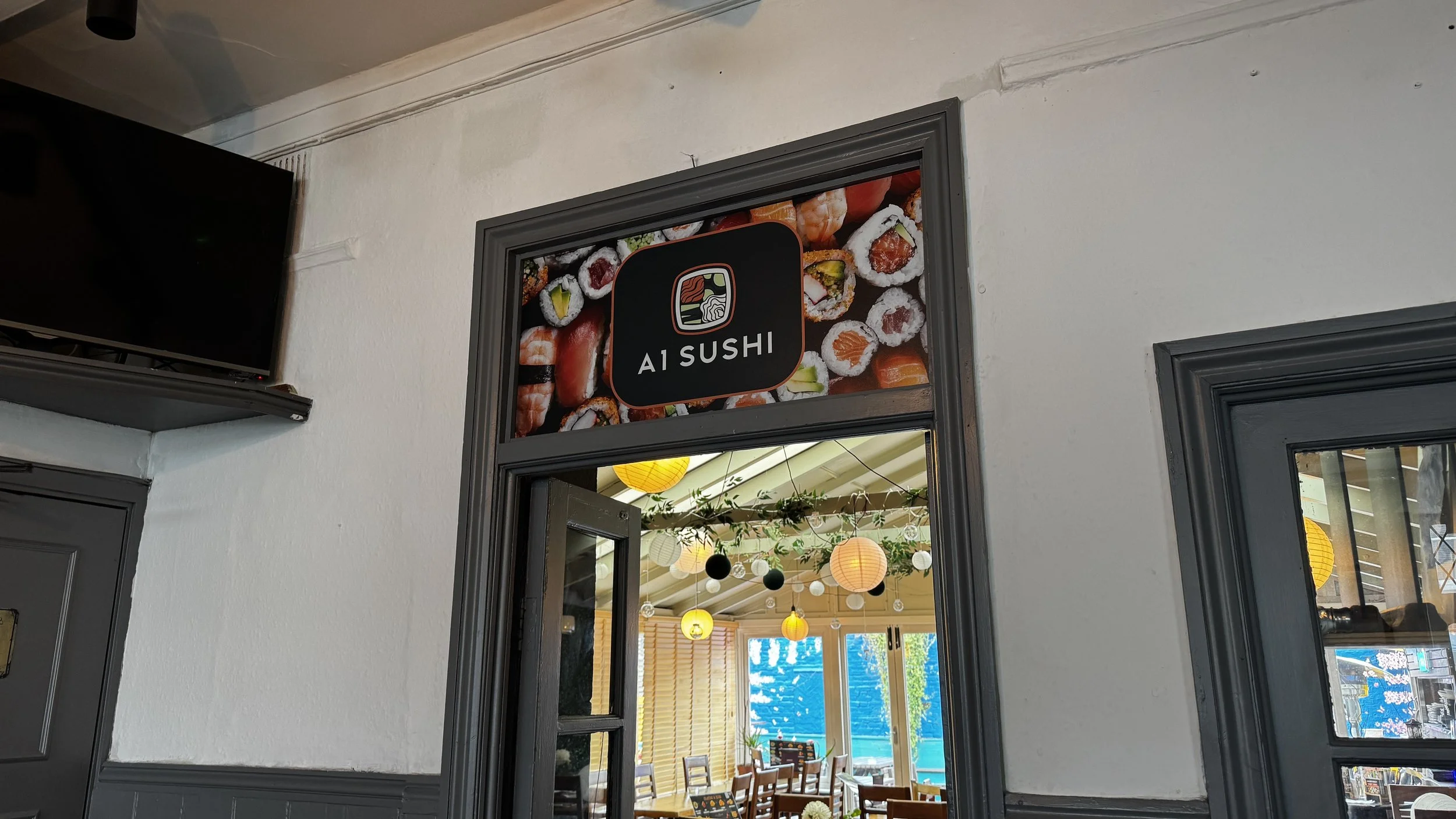 Sign reading 'A1 Sushi' with images of sushi rolls above the doorway of a restaurant, indoors with hanging lantern lights and large windows showing the outside.