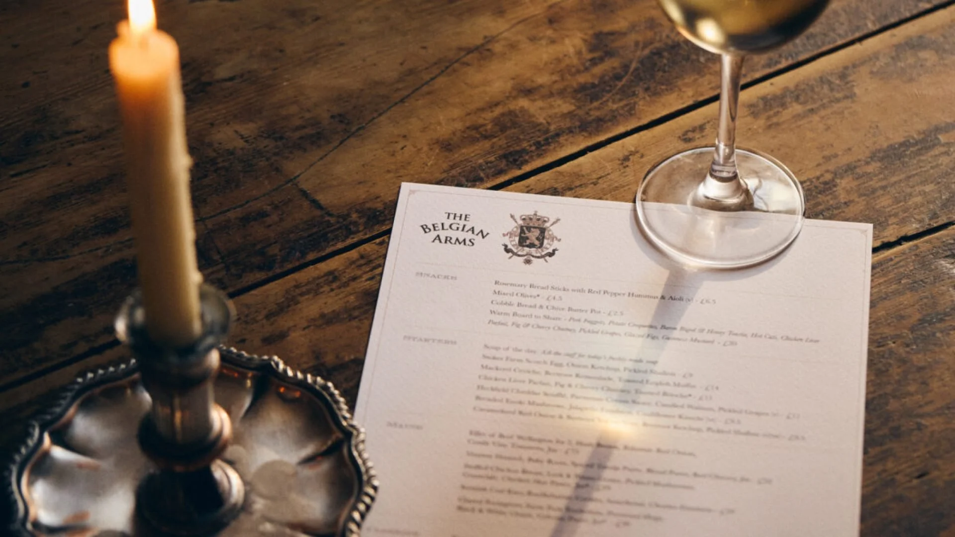 A candle, a wine glass, and a menu titled 'The Belgian Arms' on a wooden table.