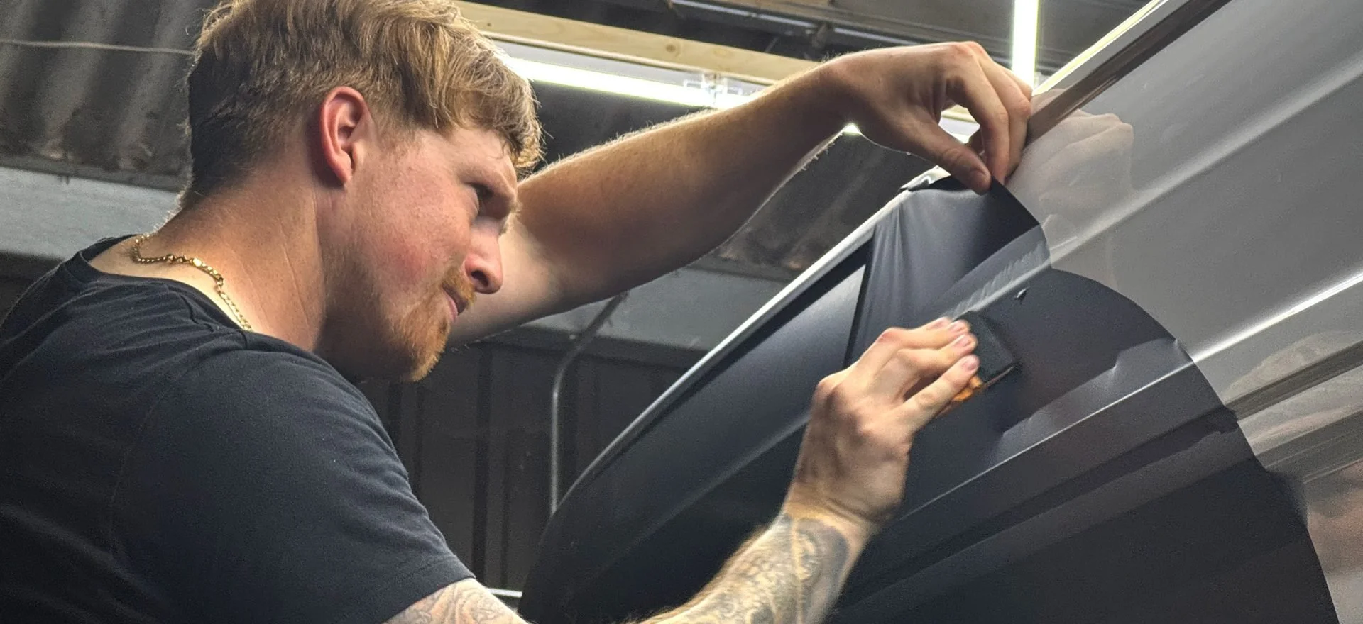 A man with reddish hair and tattoos on his arm is working on a vehicle, applying or removing a film or decal on the car's surface.