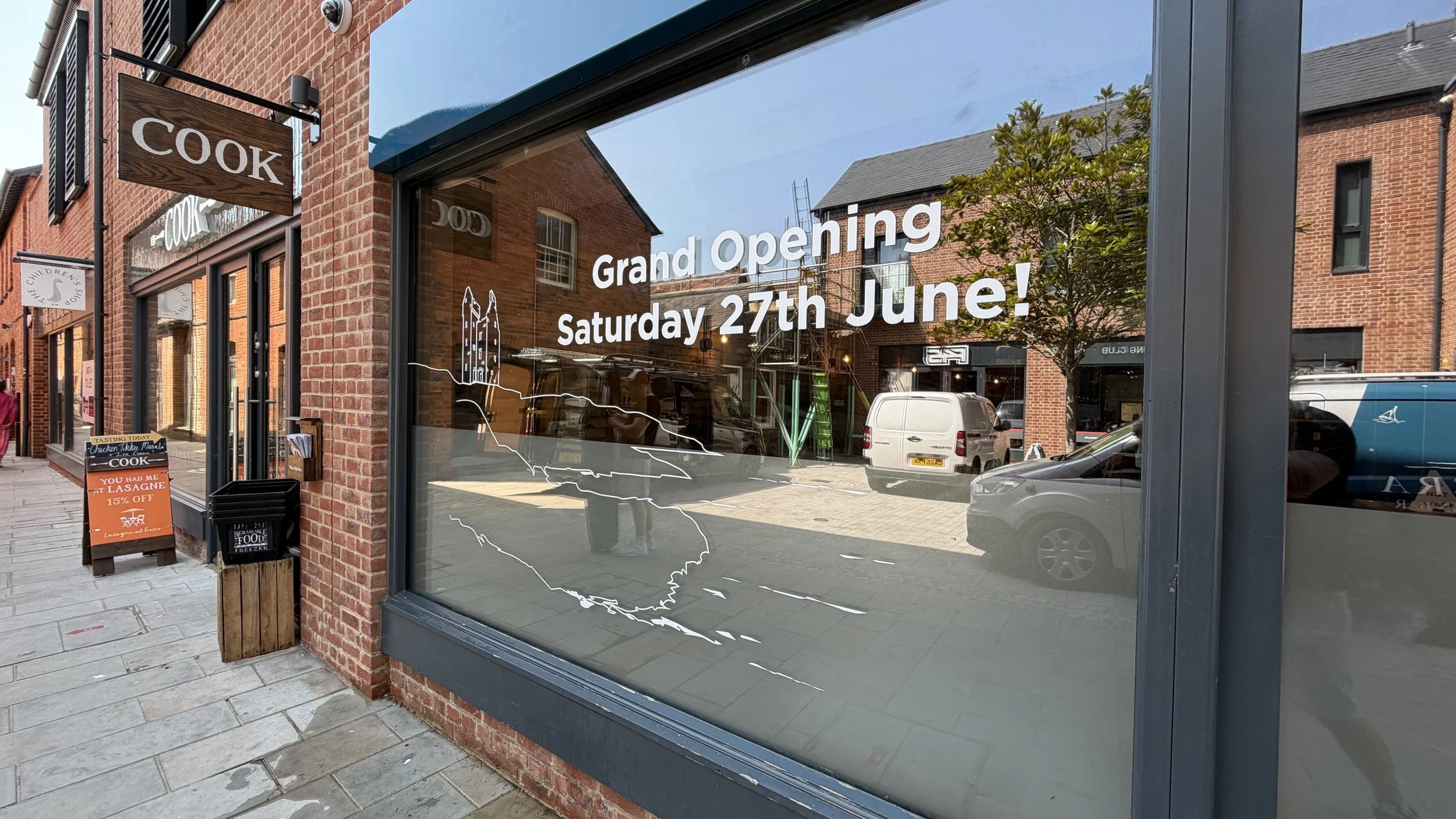 Storefront window with a sign announcing a grand opening on Saturday, June 27th, and a smaller sign outside promoting chicken tikka masala with a discount.